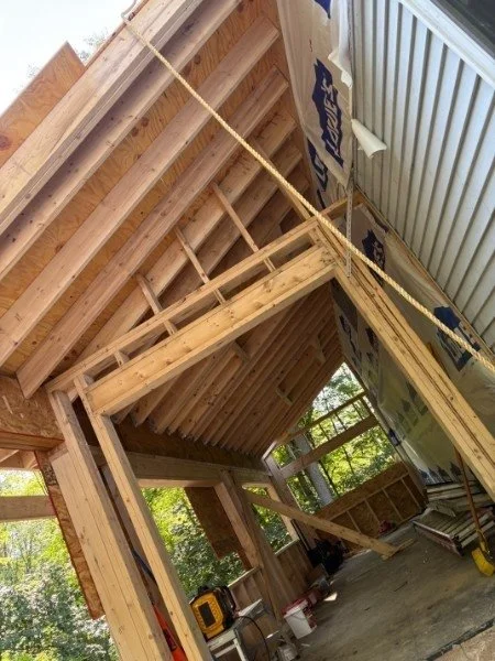 Construction site with wooden framing and insulation, partially built wall structure, and exterior view through windows showing trees.