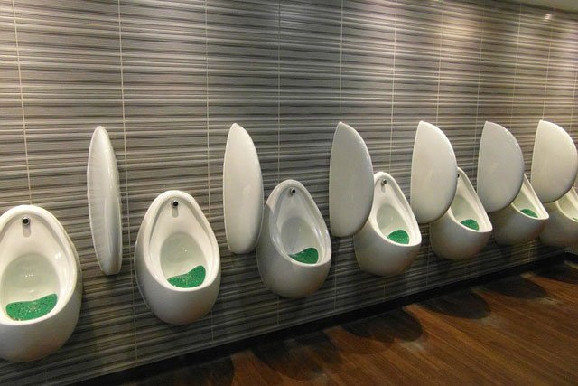 Row of six urinals with lids open, mounted on a tiled wall in a bathroom.
