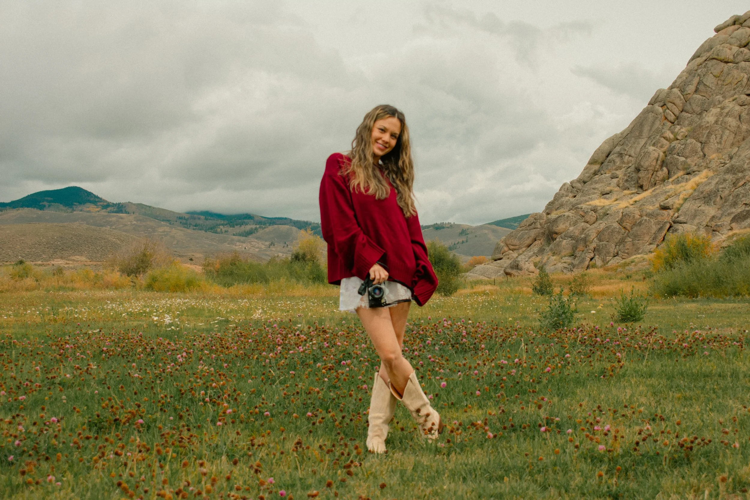 A woman with long curly hair wearing a red coat, white shorts, and tan boots stands in a field of pink and orange flowers, holding a camera. She is smiling and looking at the camera against a backdrop of rolling hills and cloudy sky.
