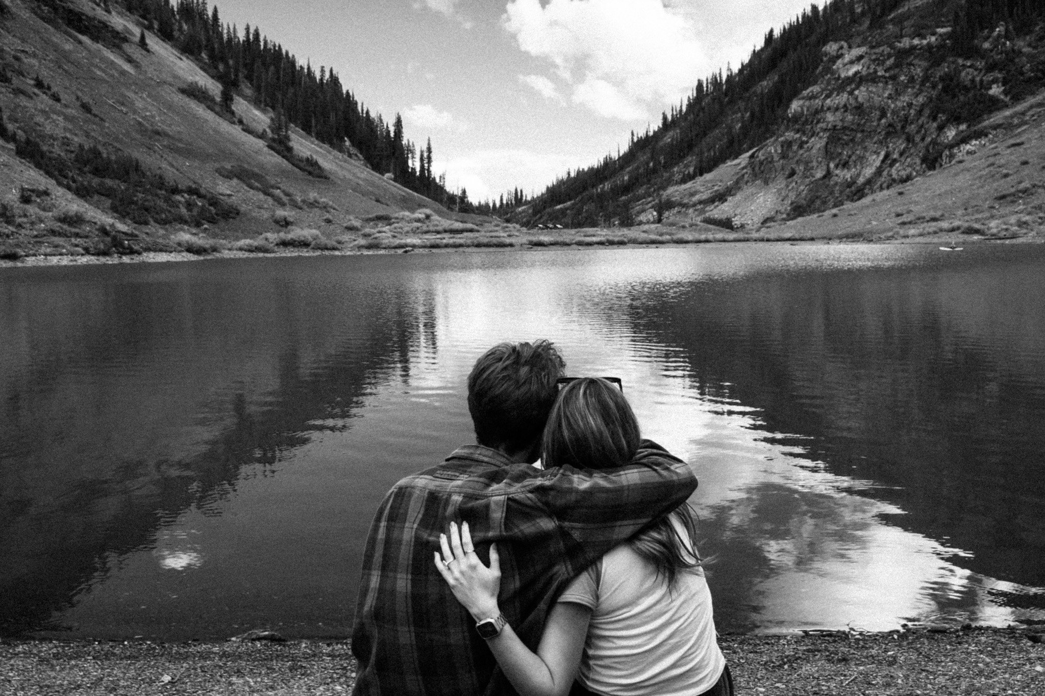 A black and white photo of a couple embracing and kissing by a lake in a mountainous area, with trees and hills in the background.