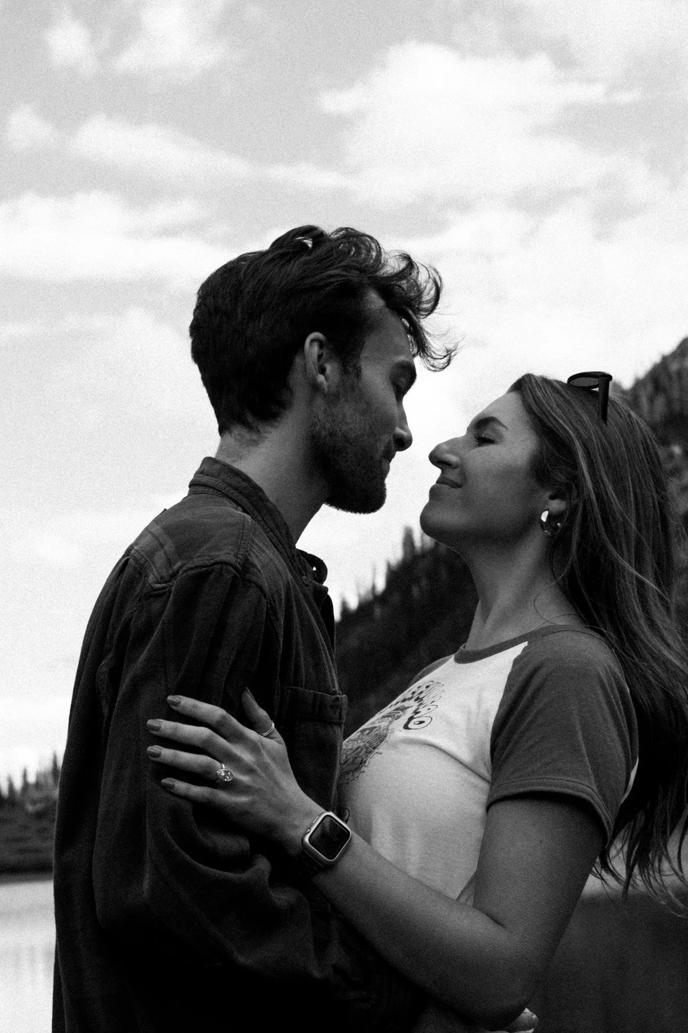 A black and white photo of a couple standing close together, smiling with their foreheads touching, outdoors with trees and sky in the background.