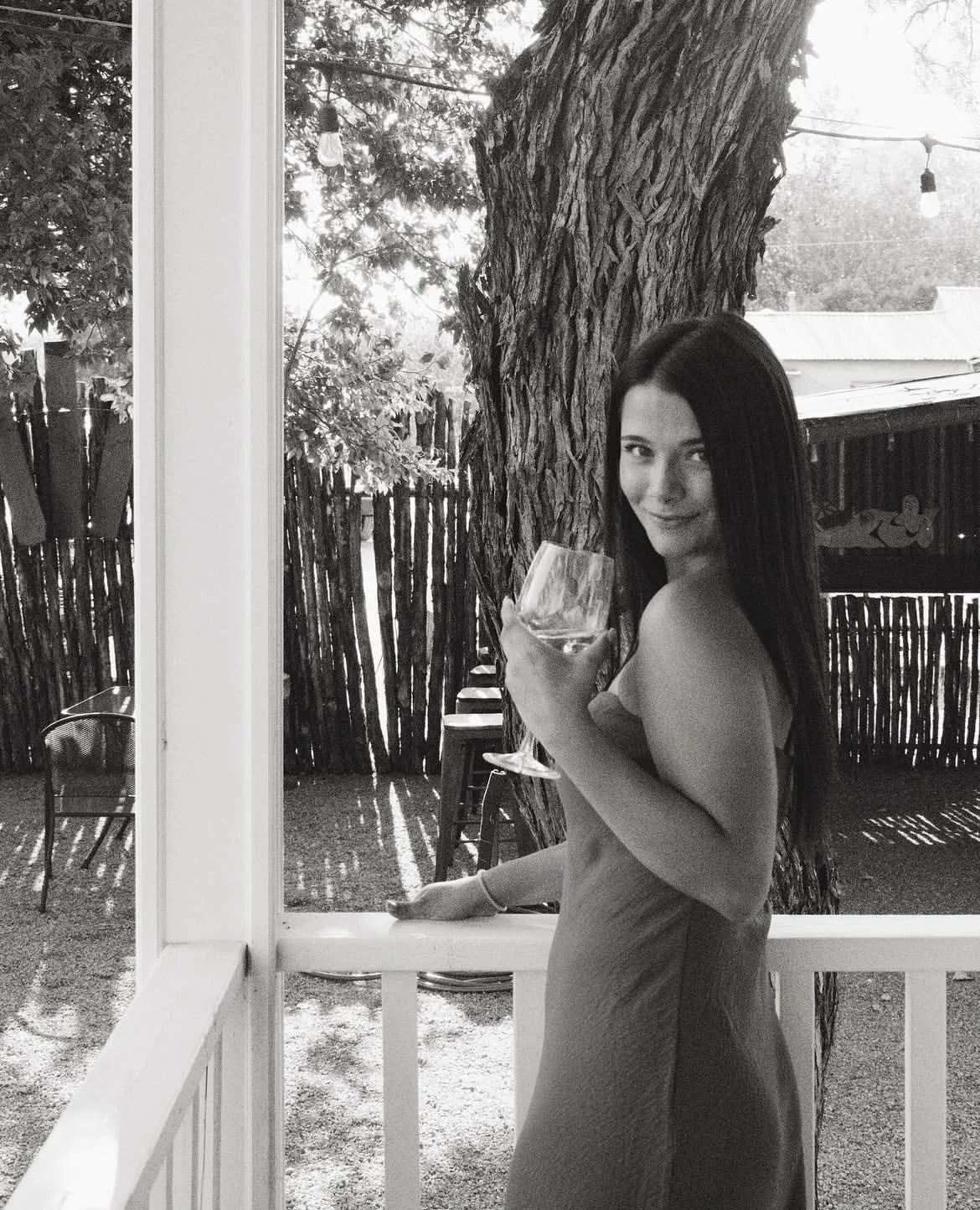 A woman with long dark hair standing outdoors near a tree, holding a glass of wine and smiling at the camera.