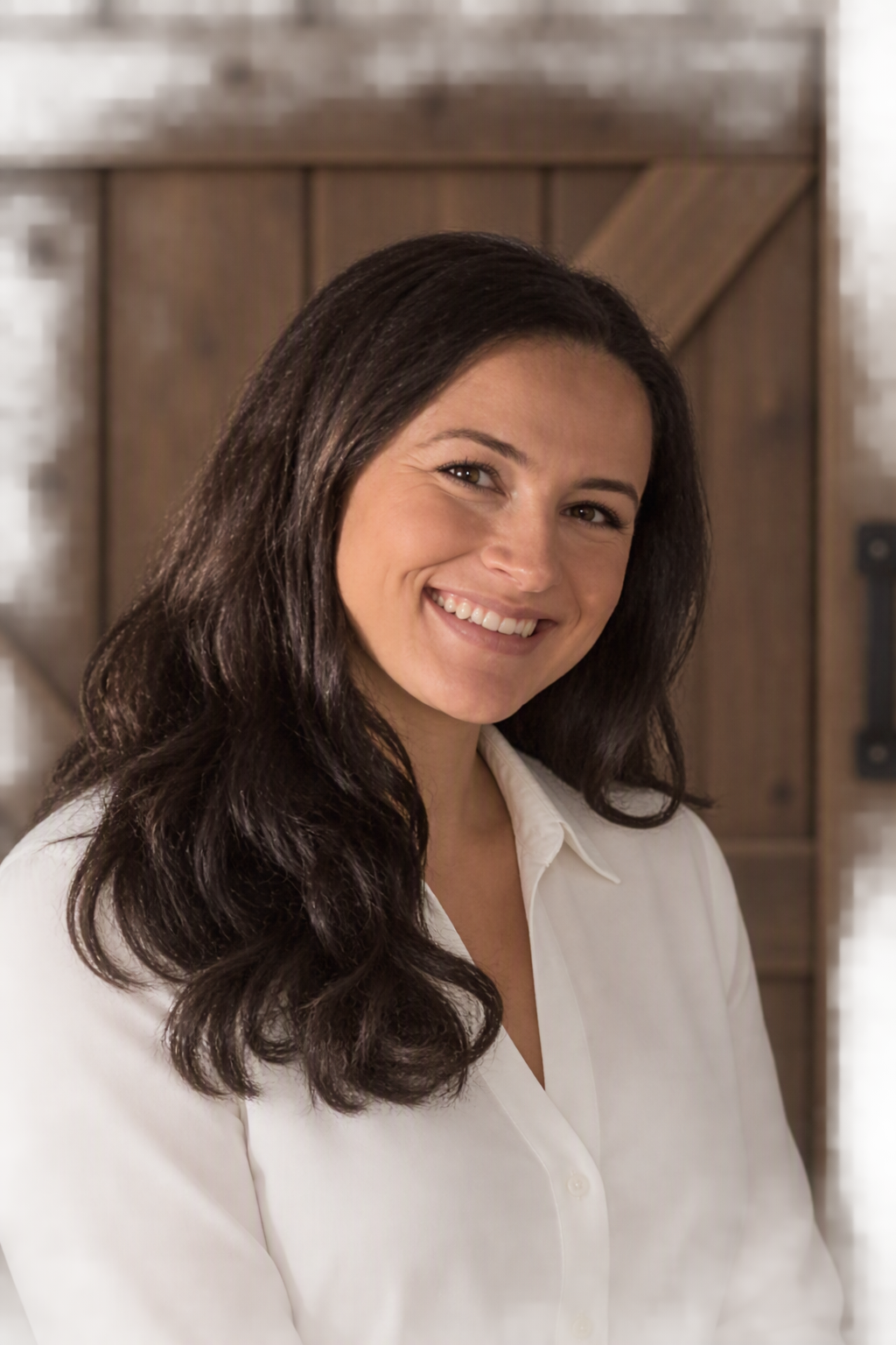 A woman with long dark hair in a white blouse, smiling, in front of a wooden background.