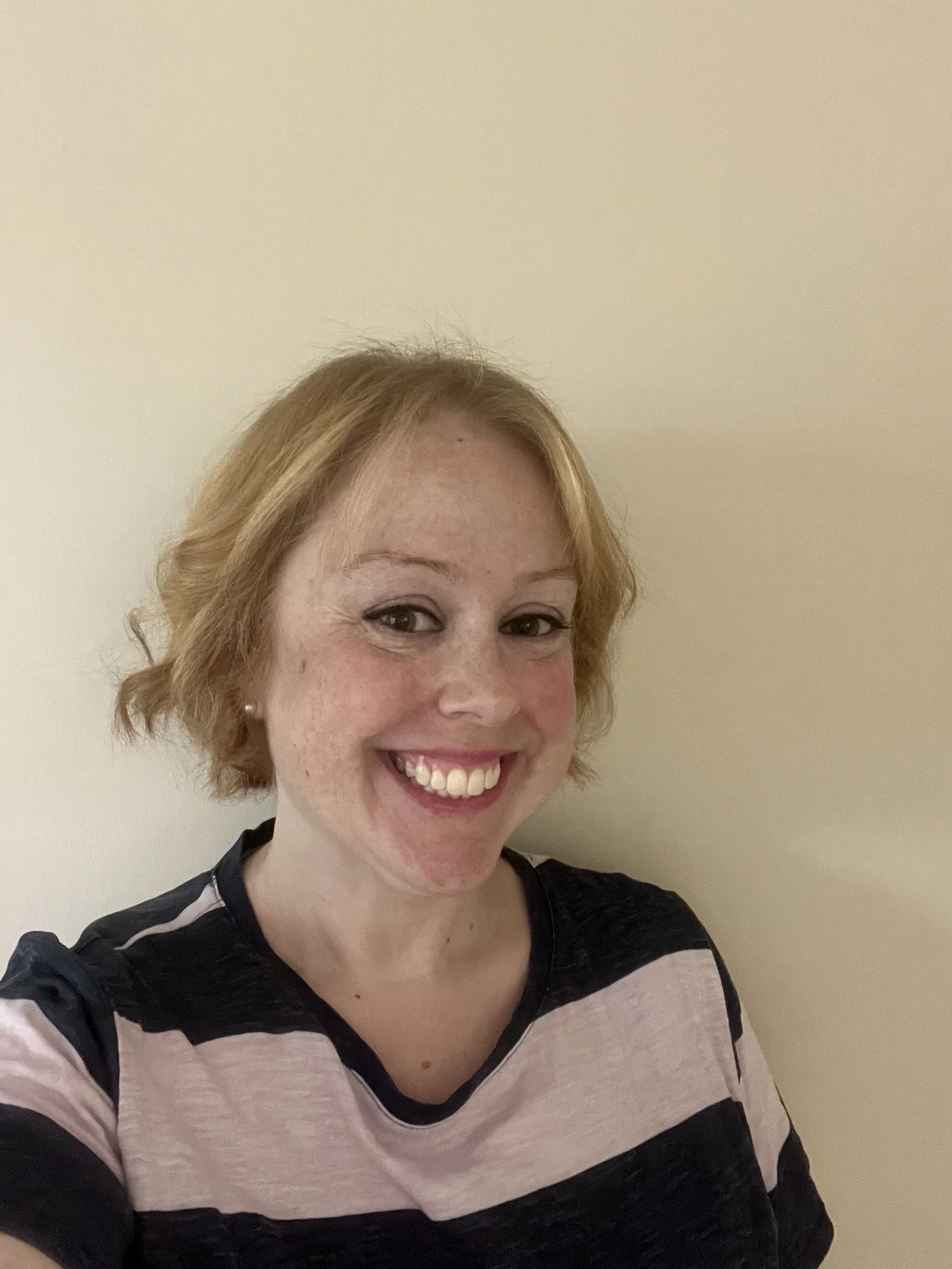 Smiling woman with blonde hair and freckles, wearing a black and pink striped shirt, standing against a plain beige wall.