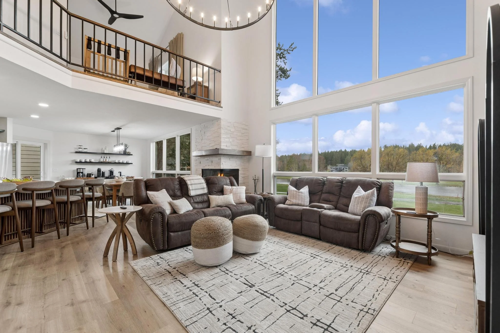 Living room with large windows showing trees and sky outside, brown leather sofas, a patterned rug, small side tables, and a cozy fireplace, with a high ceiling and modern decor.