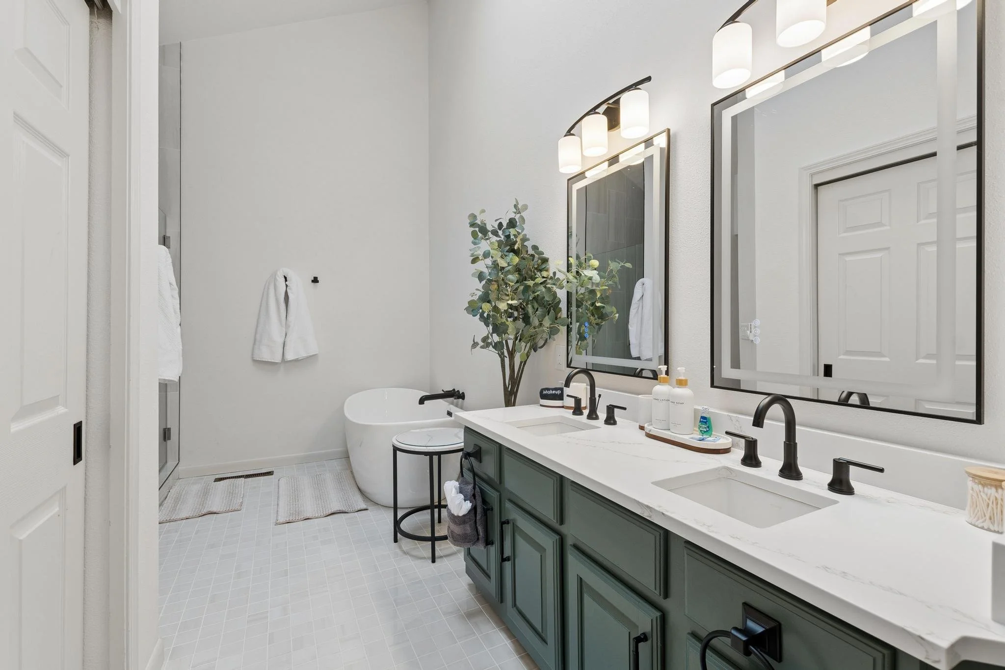 Modern bathroom with double vanity, large mirror, black fixtures, a freestanding bathtub, and decorative plants, illuminated by modern light fixtures.