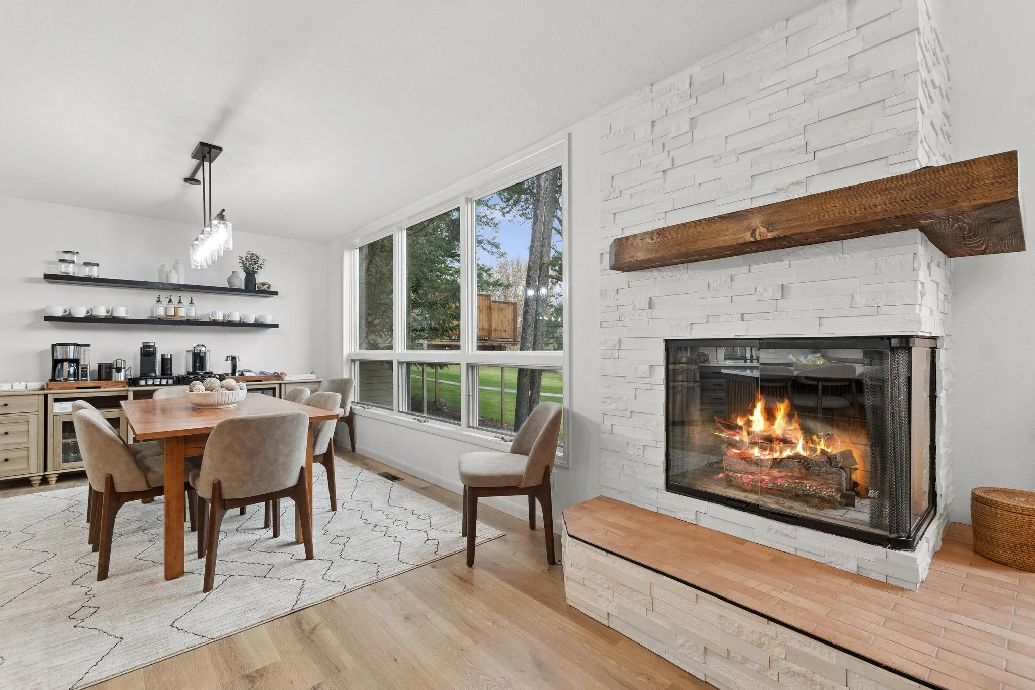 Open-concept living and dining area with a white brick fireplace, wooden mantle, a fire burning, large windows showing trees outside, a wooden dining table with beige upholstered chairs, wall-mounted shelves with dishes and decorative items, and a ce