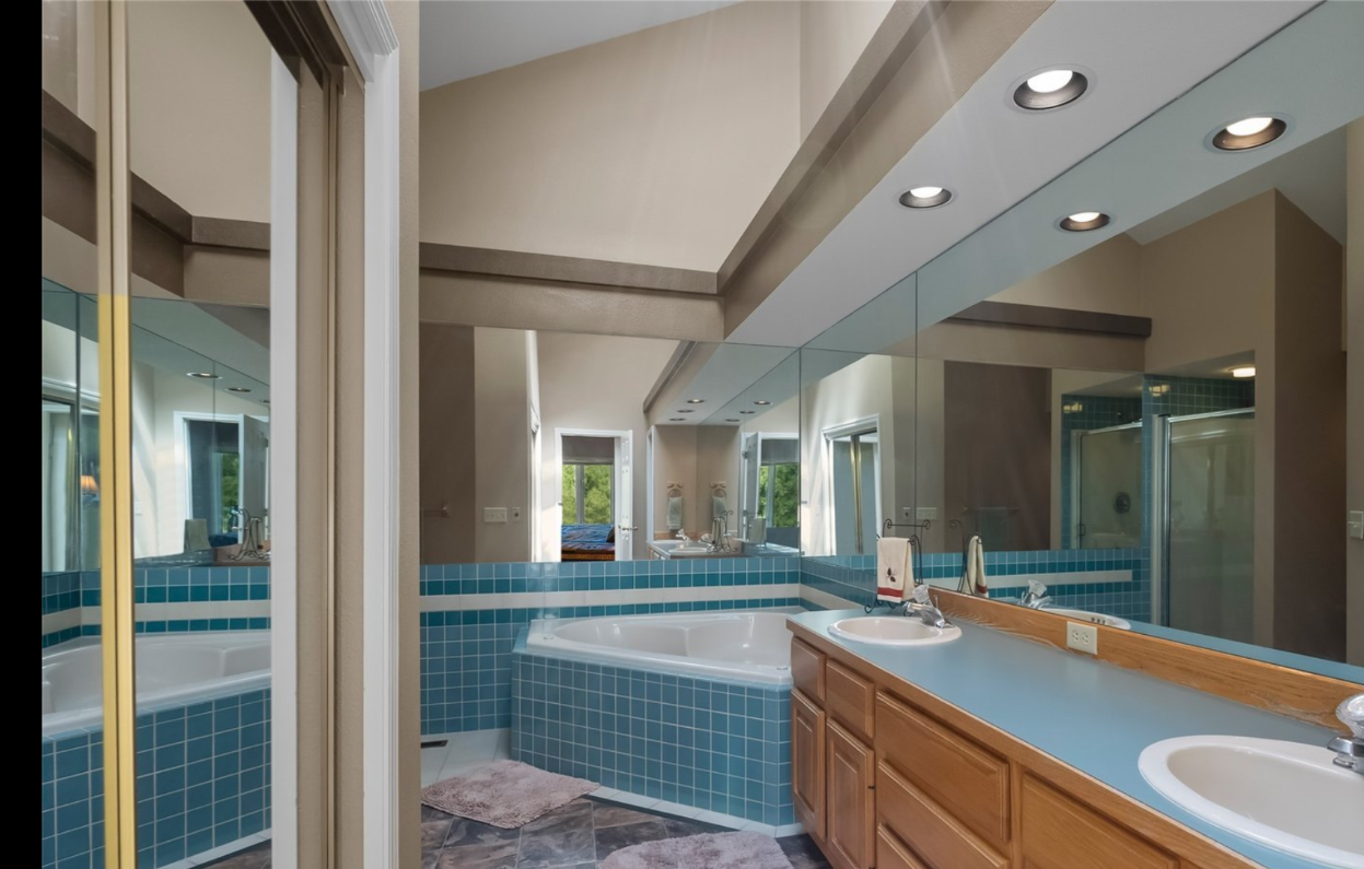 Bathroom with a large mirror, double sink vanity, corner bathtub with blue tile, and reflection of windows and a shower stall.