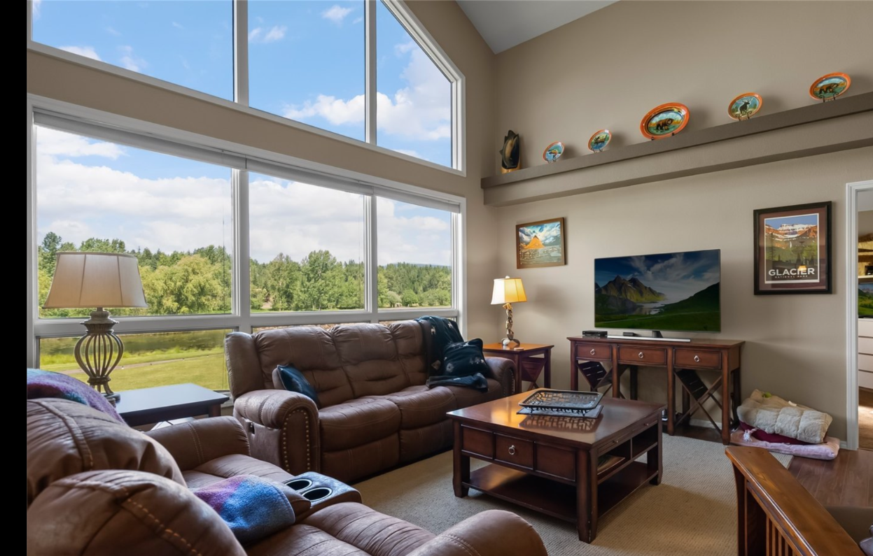Living room with large windows, brown leather couches, a wooden coffee table, a TV on a wooden stand, framed artwork, and decorative plates displayed on a high shelf.
