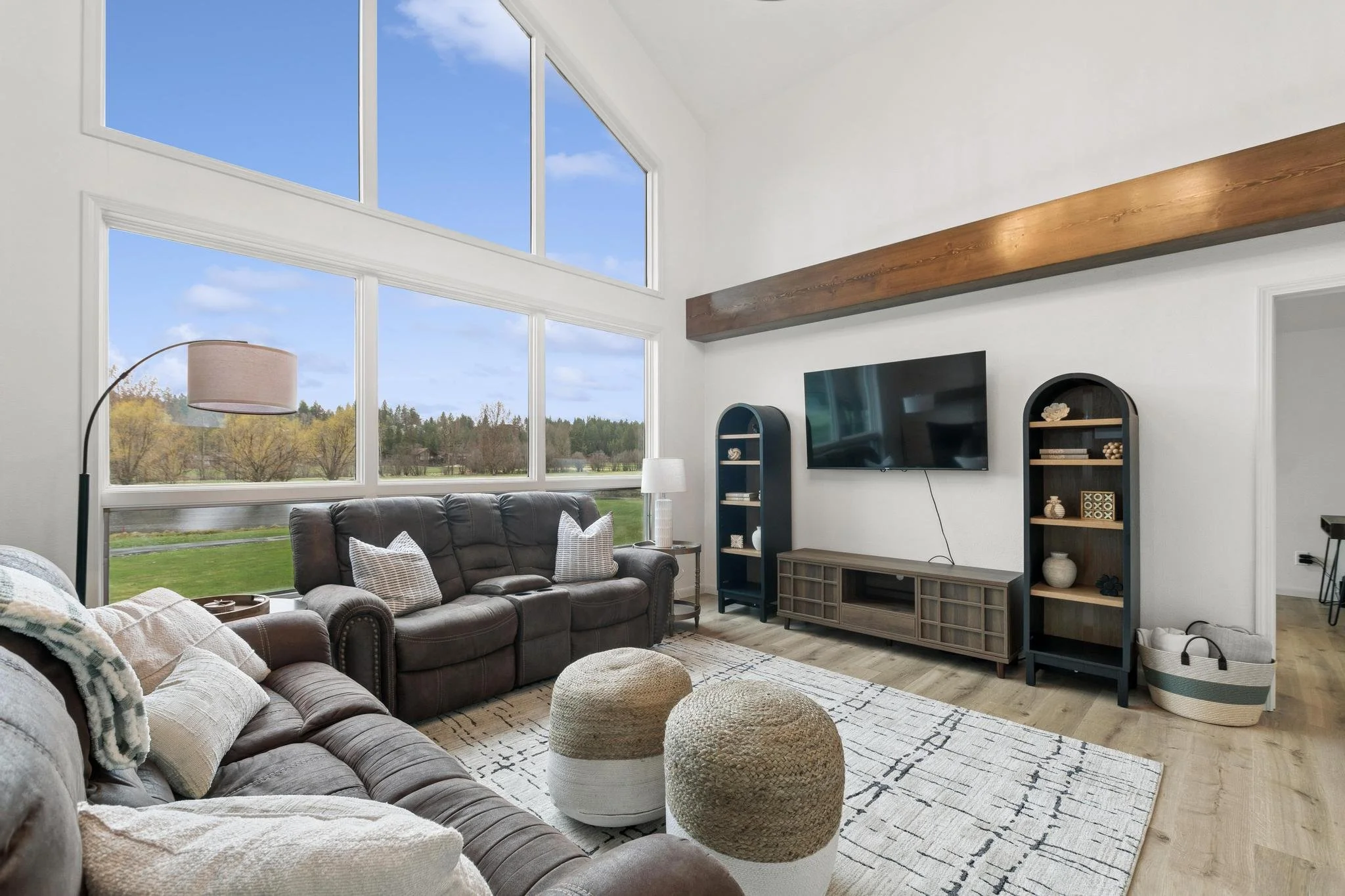 Living room with large window showing a green outdoor landscape and blue sky, featuring a dark brown leather sofa, a beige armchair, two woven ottomans, a standing lamp, and a wall-mounted TV with black shelves.