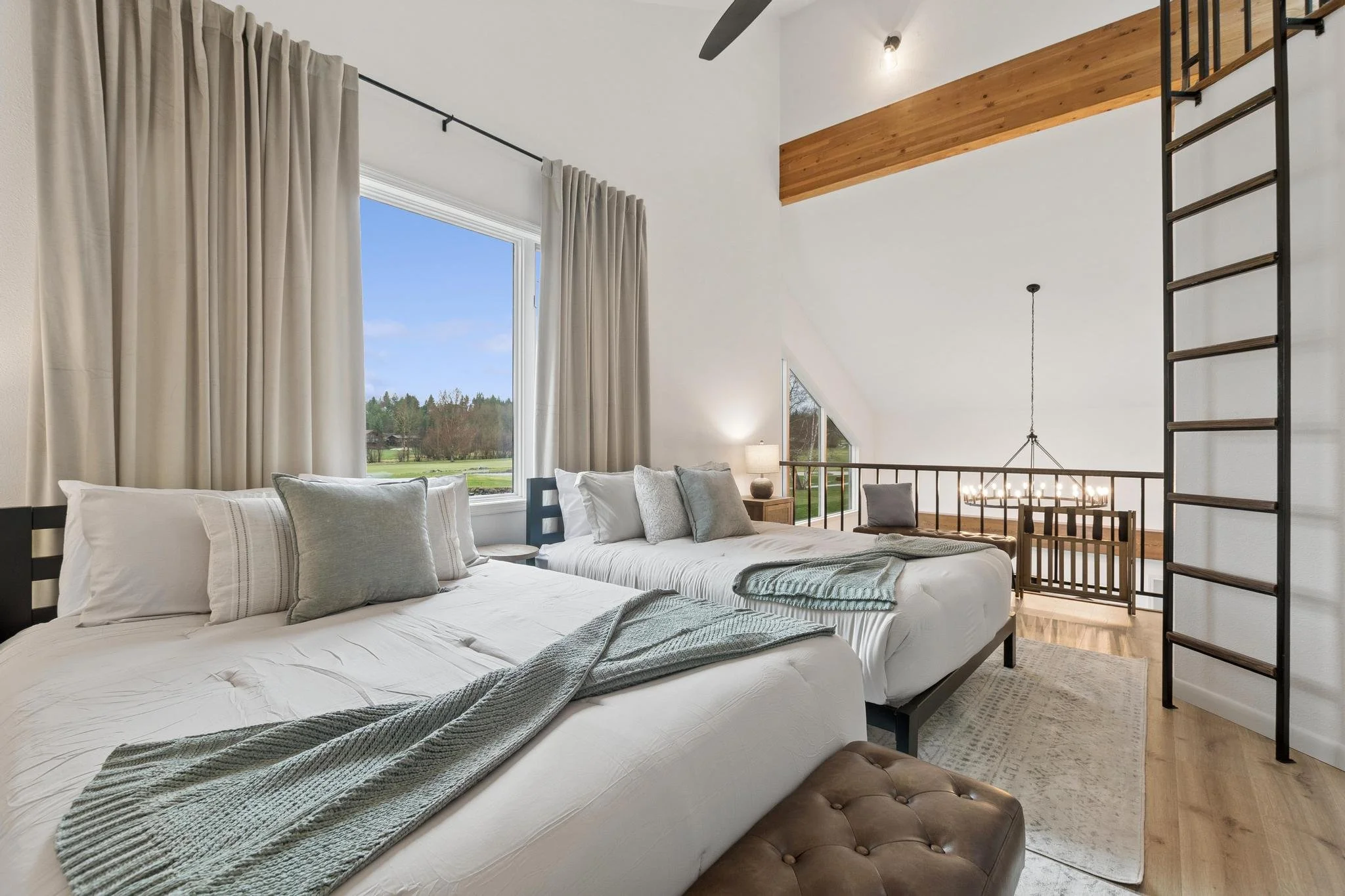 A modern bedroom with large windows, beige curtains, and a view of greenery outside. The room features white walls, a bed with multiple pillows, a bedside table with a lamp, a ceiling fan, a wooden accent wall, and a metal ladder leading to a loft ar