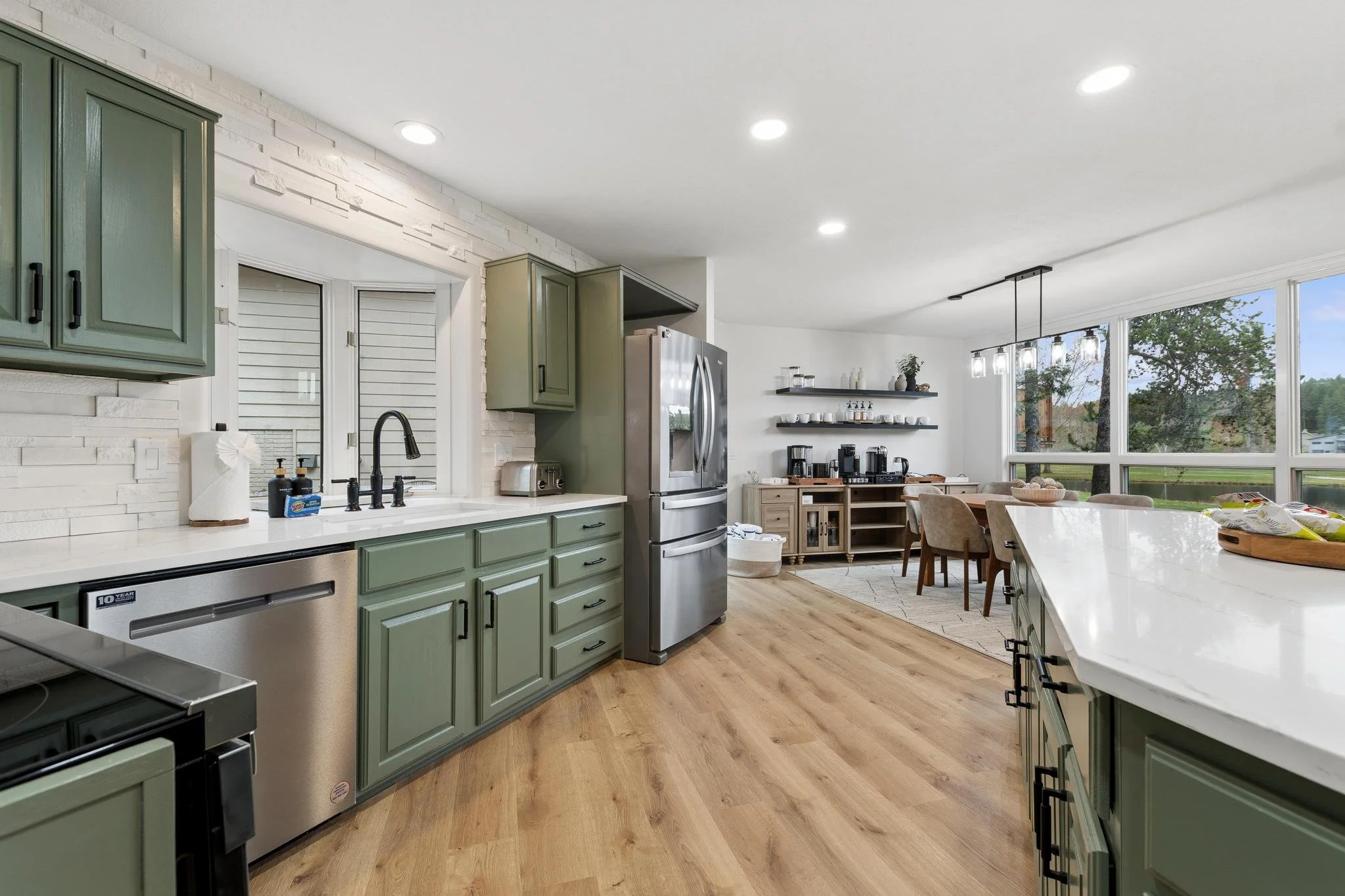 A modern kitchen with green cabinets, stainless steel refrigerator, white countertops, light wood flooring, and large windows showing trees and blue sky outside.