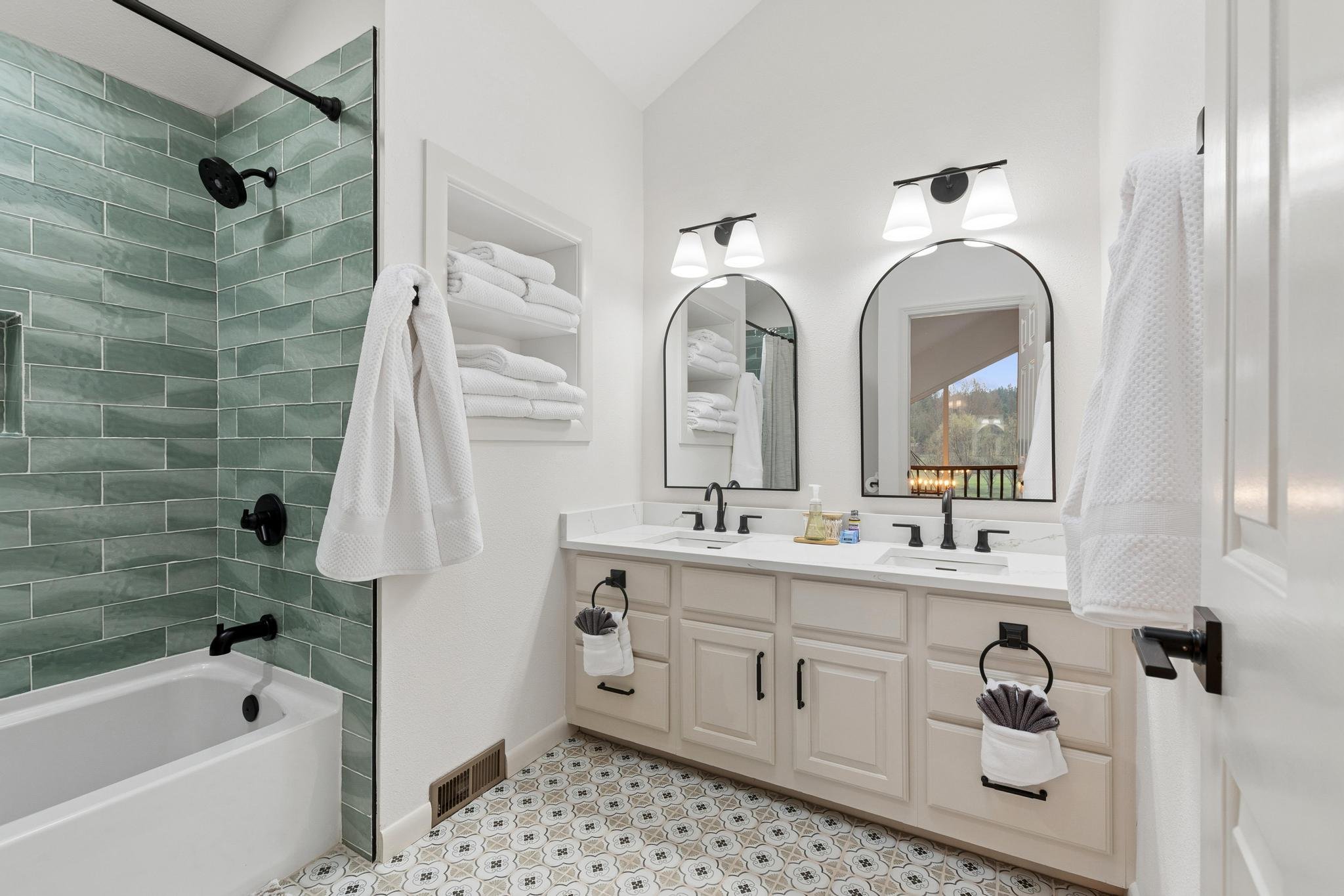 Bathroom with a walk-in shower with green tiles, white walls, a double vanity with two mirrors, and patterned floor tiles.