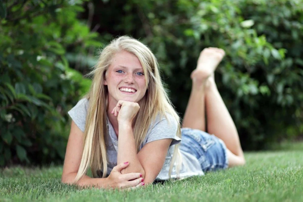 A young woman with long blonde hair lying on grass in a garden, smiling and resting her chin on her hand.