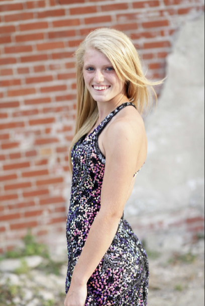A young woman with long blonde hair smiling, wearing a sparkly floral dress, standing outdoors in front of a brick wall.
