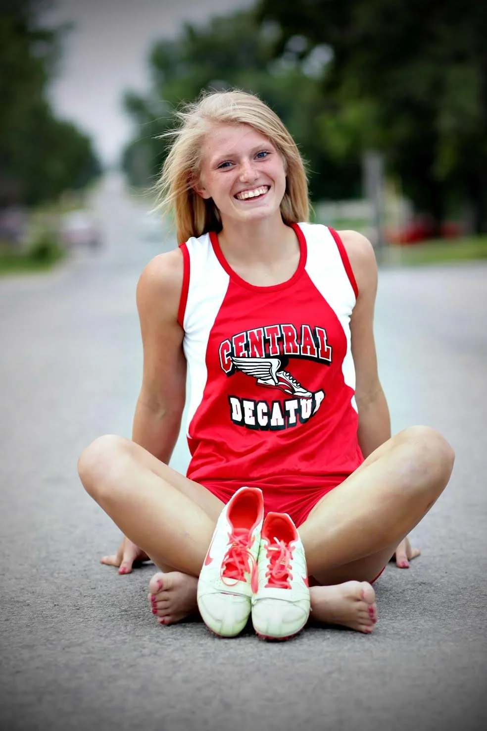 A young girl with blonde hair, wearing a red and white athletic shirt with 'Central Decatur' written on it, sits cross-legged on the pavement, smiling, with her sneakers placed in front of her.