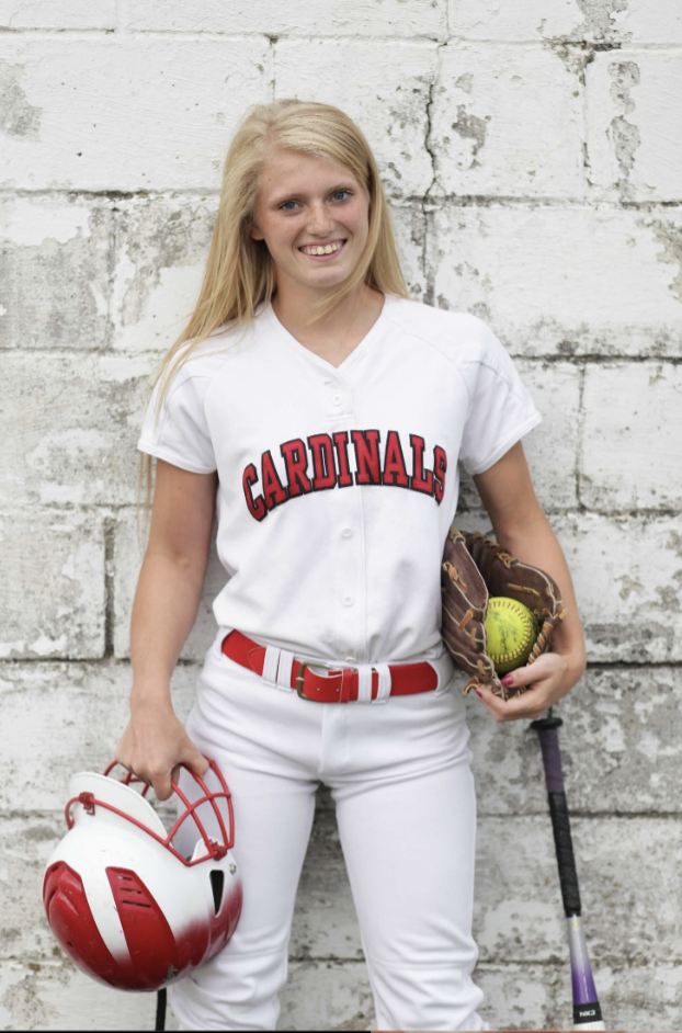 A young woman in a white baseball uniform with 'Cardinals' written on it, holding a baseball helmet and glove, standing against a white brick wall, with a baseball bat and a ball in the glove, smiling at the camera.