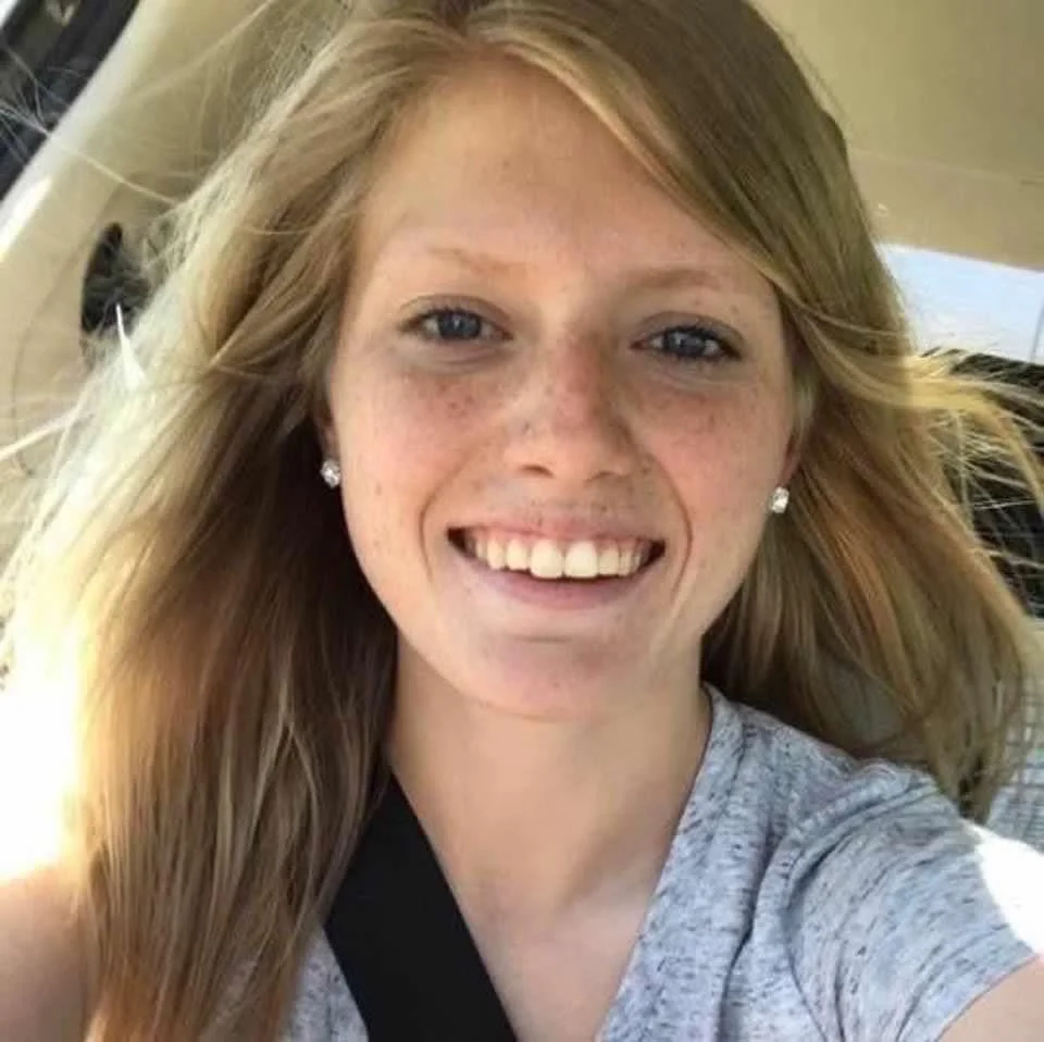 A cheerful young woman with long blonde hair, blue eyes, and freckles smiling while taking a selfie in a vehicle.