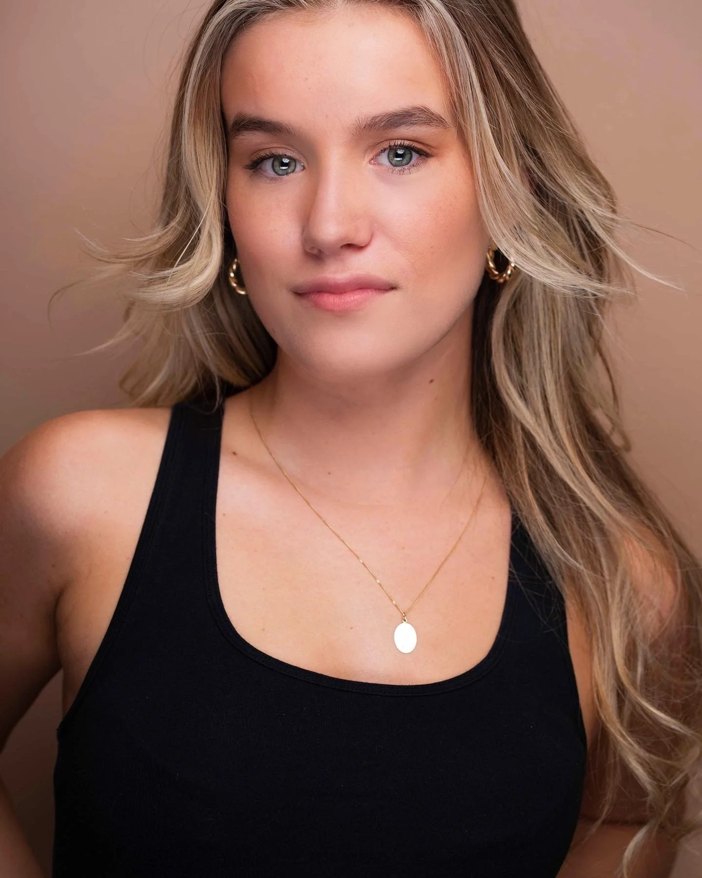 Close-up of a woman with long, wavy blonde hair, wearing a black tank top, gold hoop earrings, and a gold necklace with a small oval pendant, against a beige background.