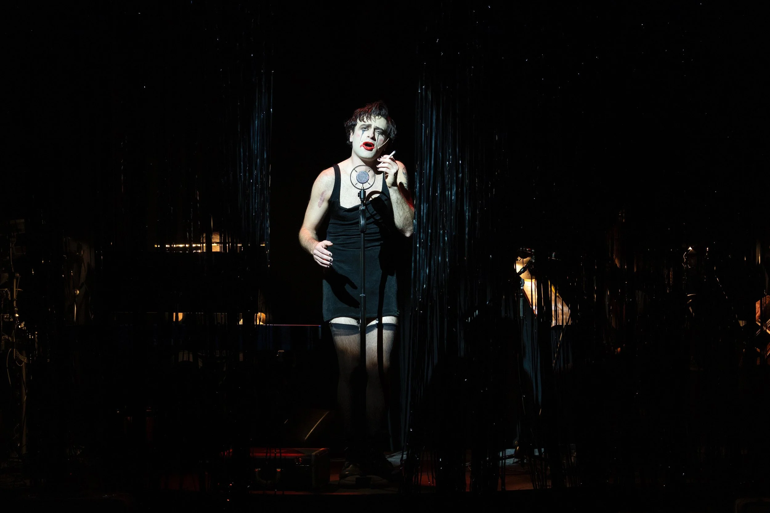 A person dressed as a clown singing into a microphone on a dark stage, with black hair, white face paint, red lipstick, black eyeshadow, wearing a black arm sleeve, a black dress, and holding a cigarette, with shiny black curtain backdrop.