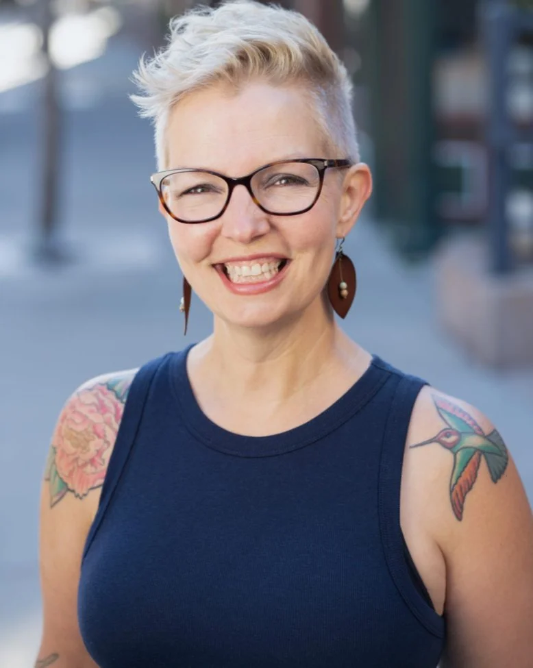 A smiling woman with short blonde hair, glasses, and tattoos on her shoulders, standing outdoors in a sunny environment.
