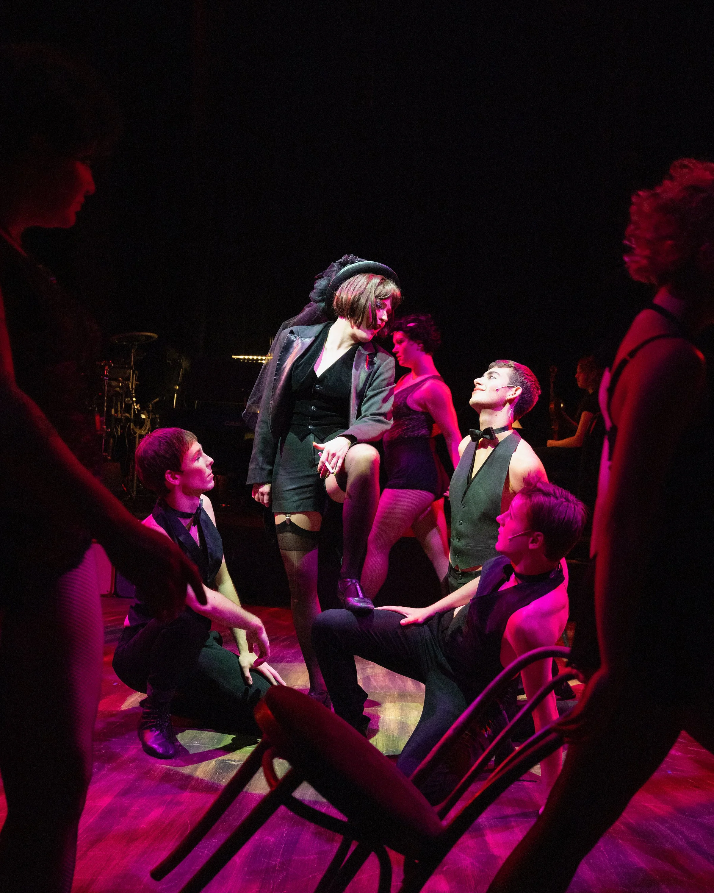 The image shows a group of performers, primarily young, in a dark setting with purple stage lighting. Some are standing while others are seated or kneeling. A woman with a bob haircut, dressed in a black blazer and skirt, appears to be engaging with 