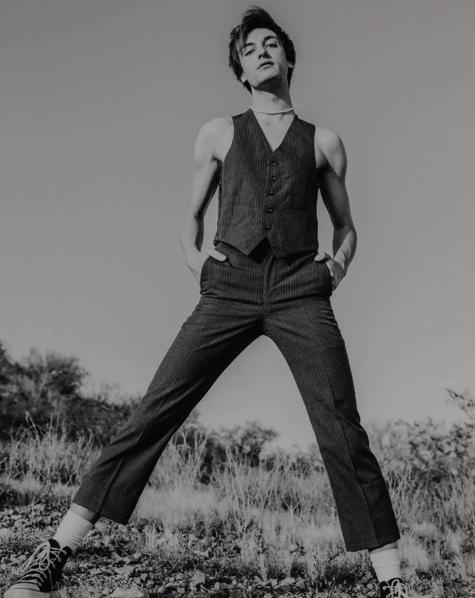 Black and white photo of a person standing outdoors on a rocky ground with plants, wearing a pinstripe vest, matching pinstripe pants, sneakers, and a choker, with hands in pockets and a confident expression.