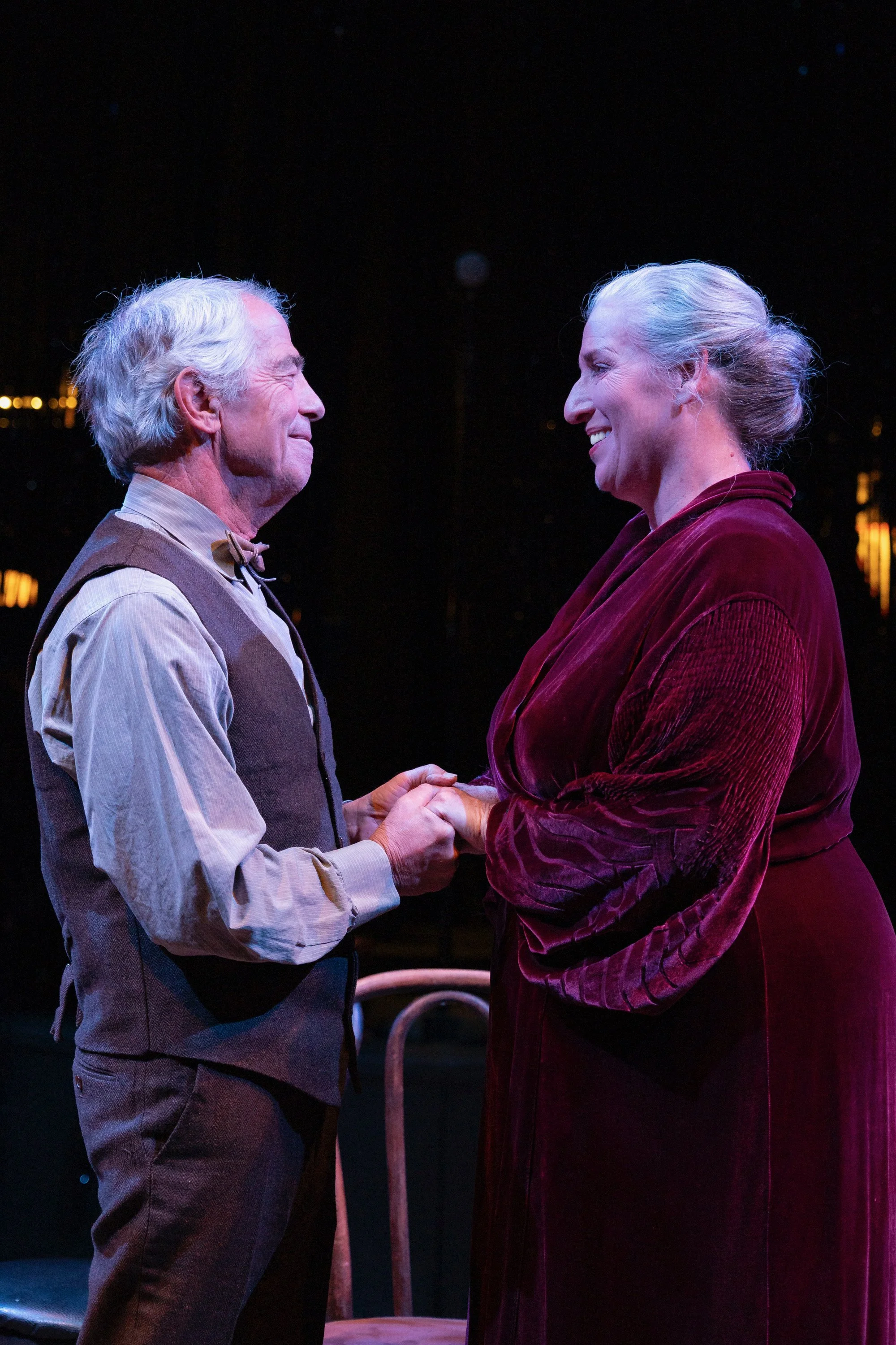 An elderly man with white hair, wearing a beige shirt, suspenders, and a bow tie, and an elderly woman with gray hair styled in an elegant bun, wearing a burgundy velvet dress, are holding hands and smiling at each other on stage at night.