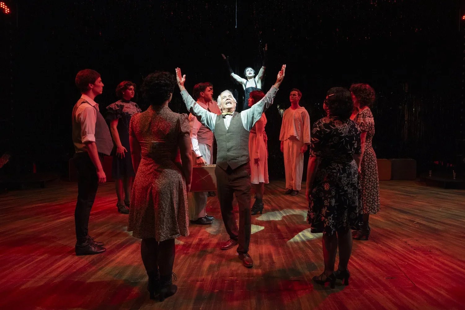 Actors performing a theatrical scene on stage with dramatic lighting, with one actor in front gesturing joyfully and others standing around them.