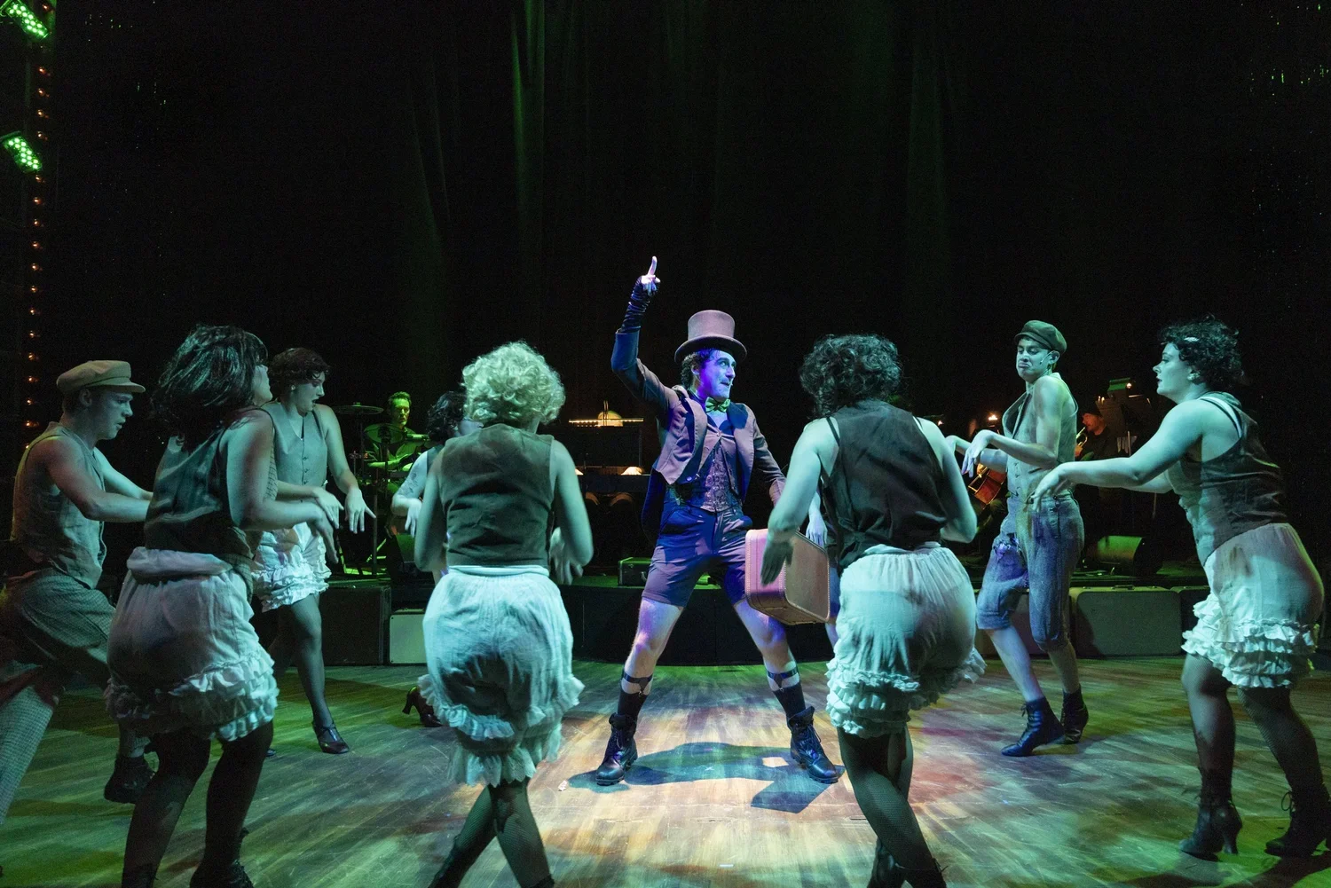 Theatrical performance with a person dressed as a ringmaster in the center, surrounded by dancers in vintage-style costumes, on a stage with dramatic lighting.