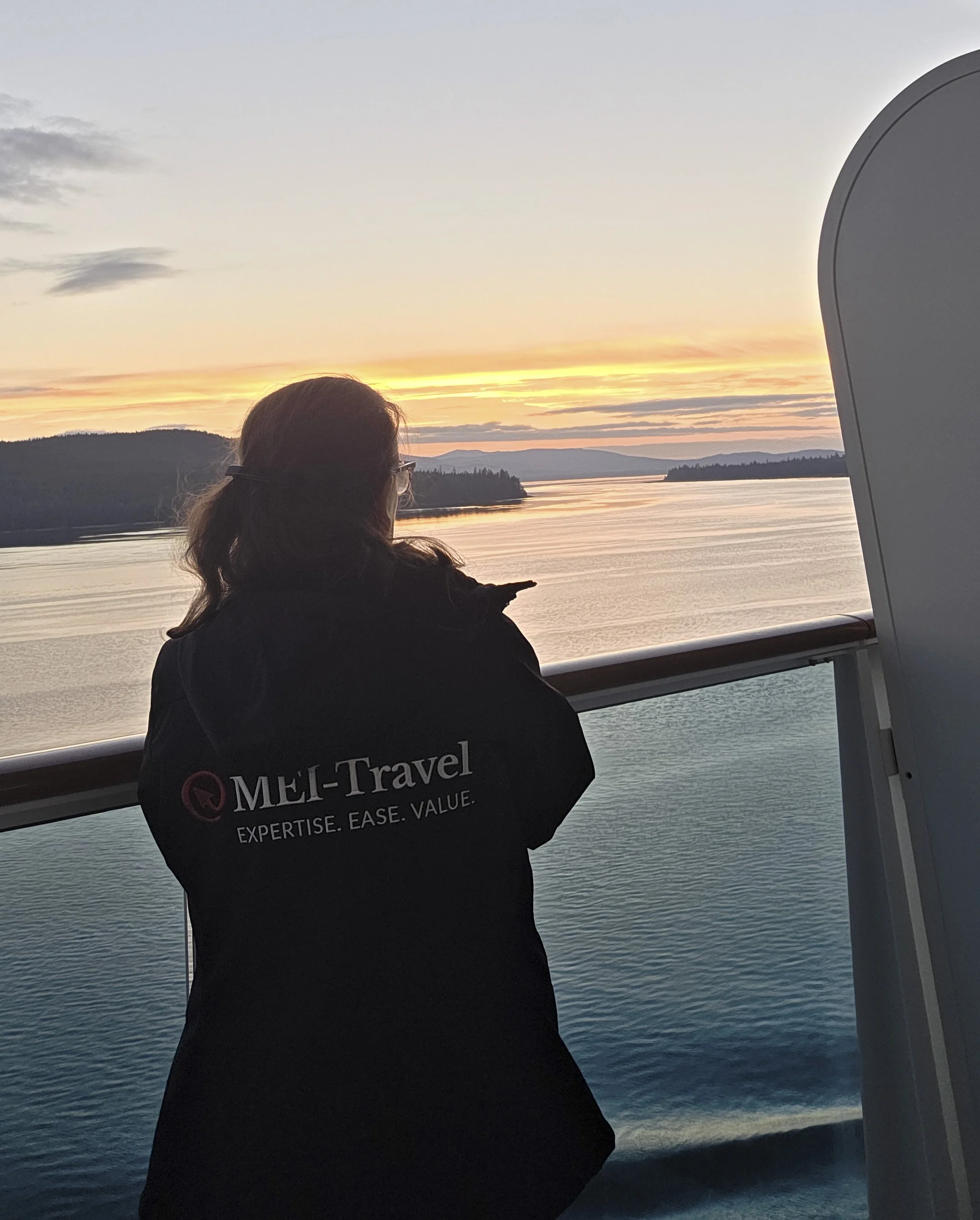 Person with long hair and glasses looking at a sunset over water from a ferry or boat, with landscape and hills in the background, and part of the boat's white structure visible.