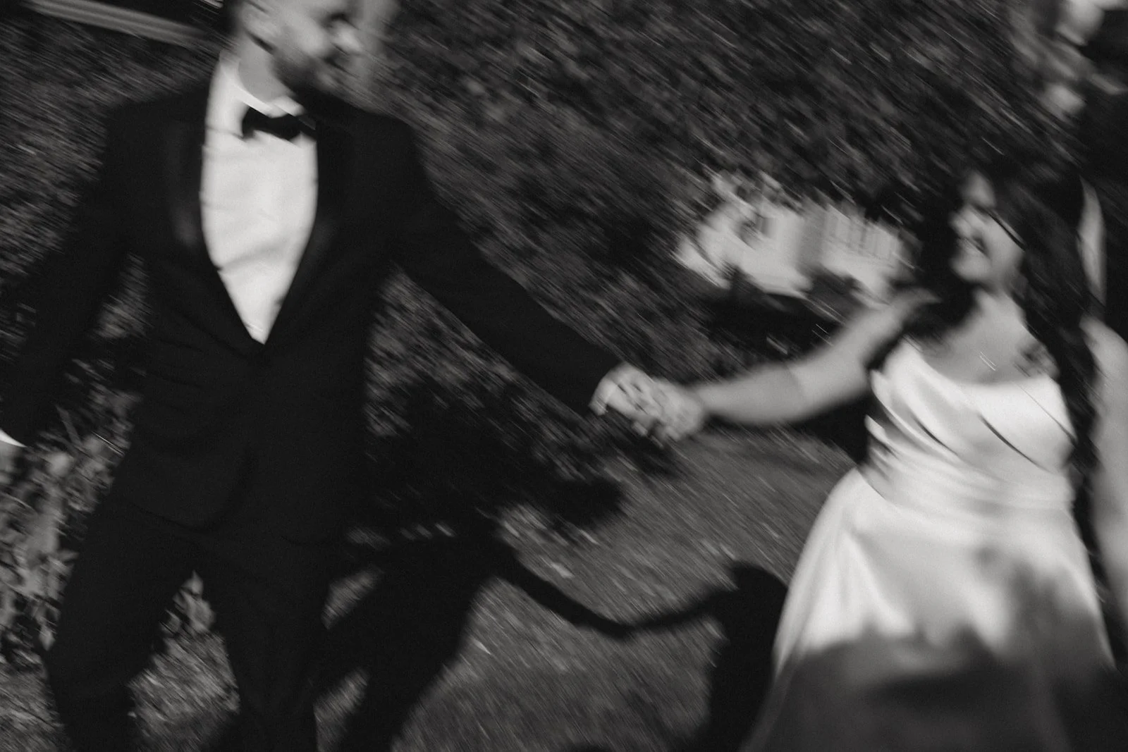 A black and white photo of a bride and groom holding hands and dancing outdoors, both dressed in formal attire.