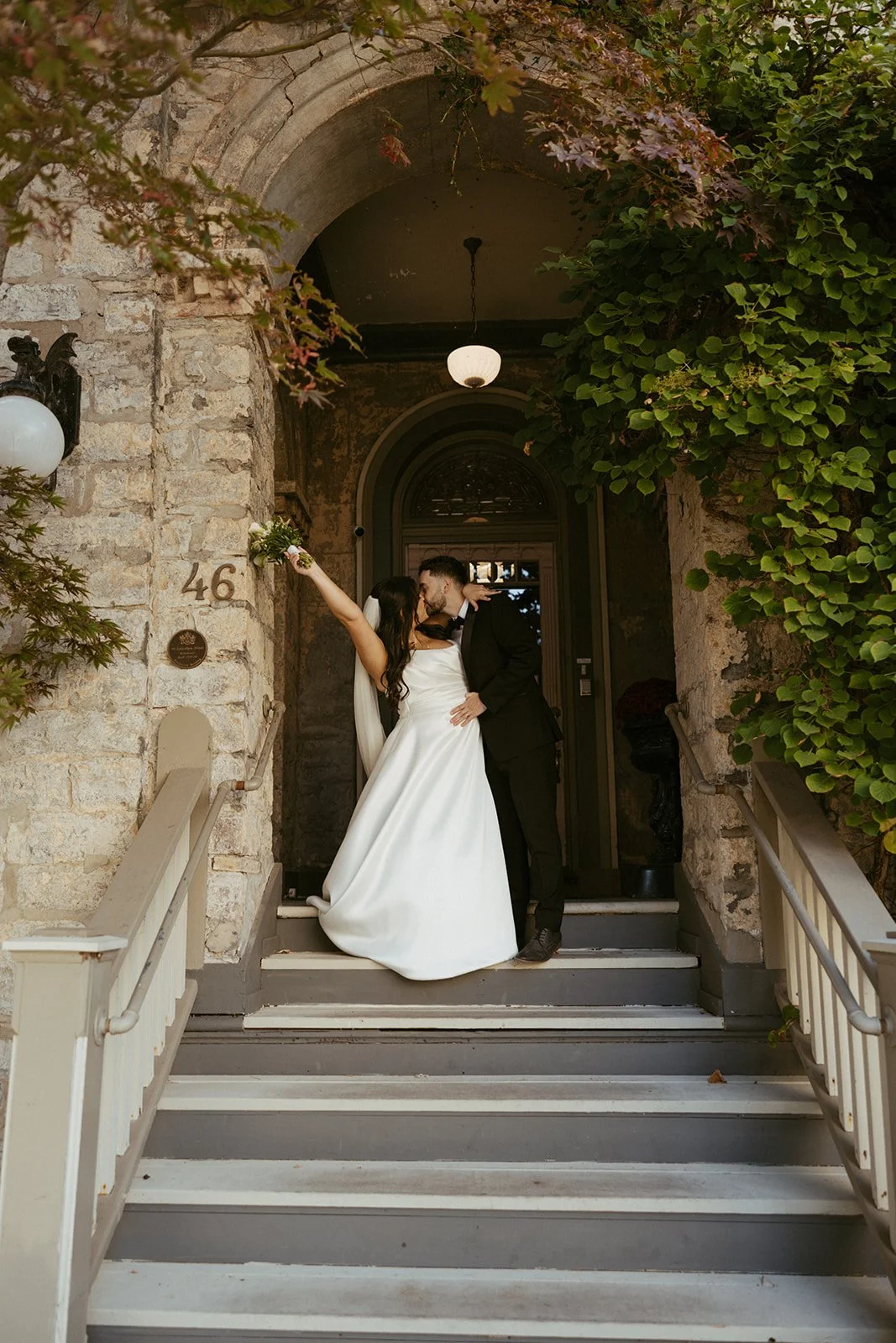 A newly married couple kissing on steps in front of a stone building with the number 46, surrounded by greenery.