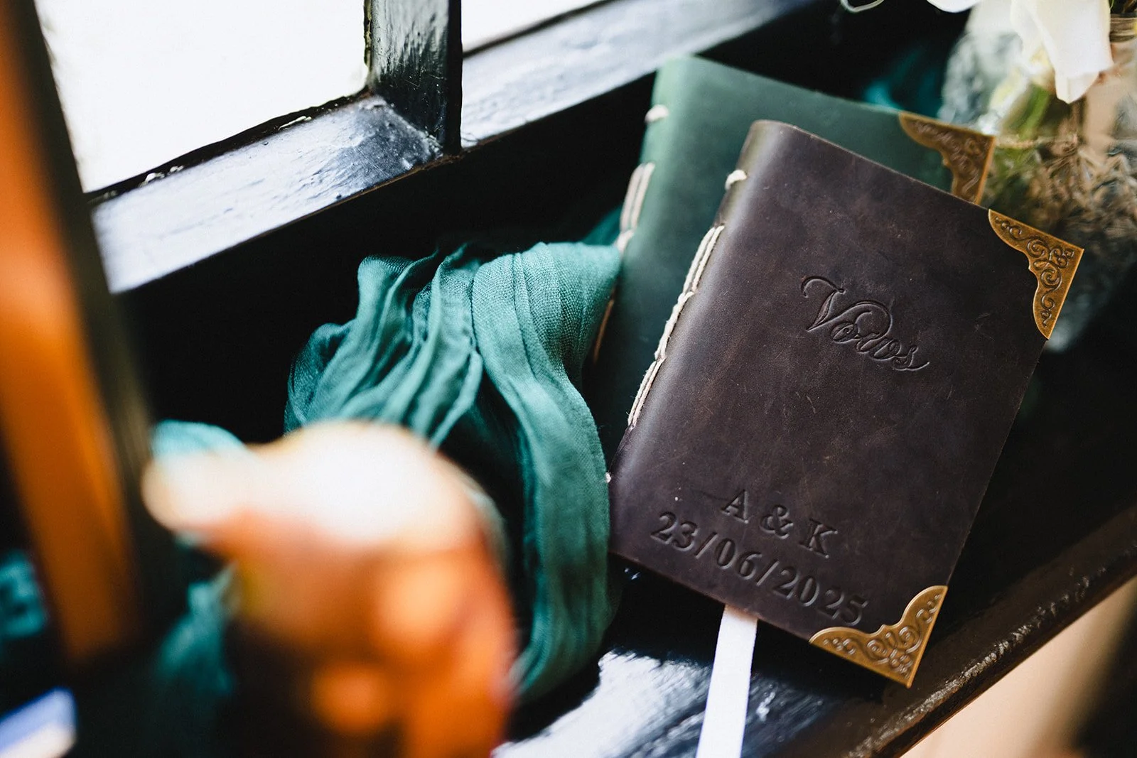 A brown leather guest book with engraved text and date, resting on a dark wooden surface, with a teal cloth and decorations nearby.