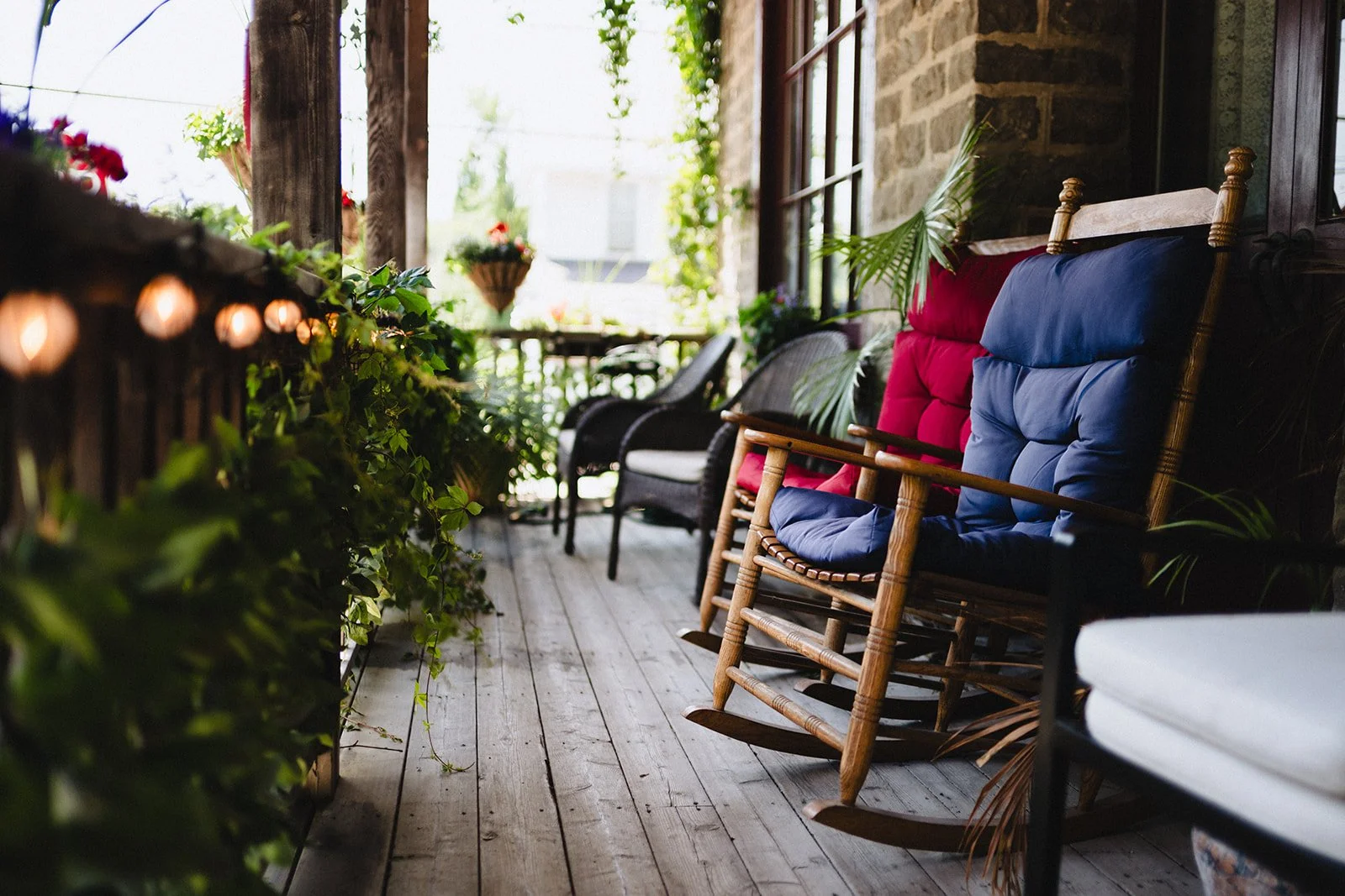 A cozy porch with wooden flooring, decorated with colorful cushions on chairs, surrounded by green plants and flowers, with sunlight filtering through the trees.