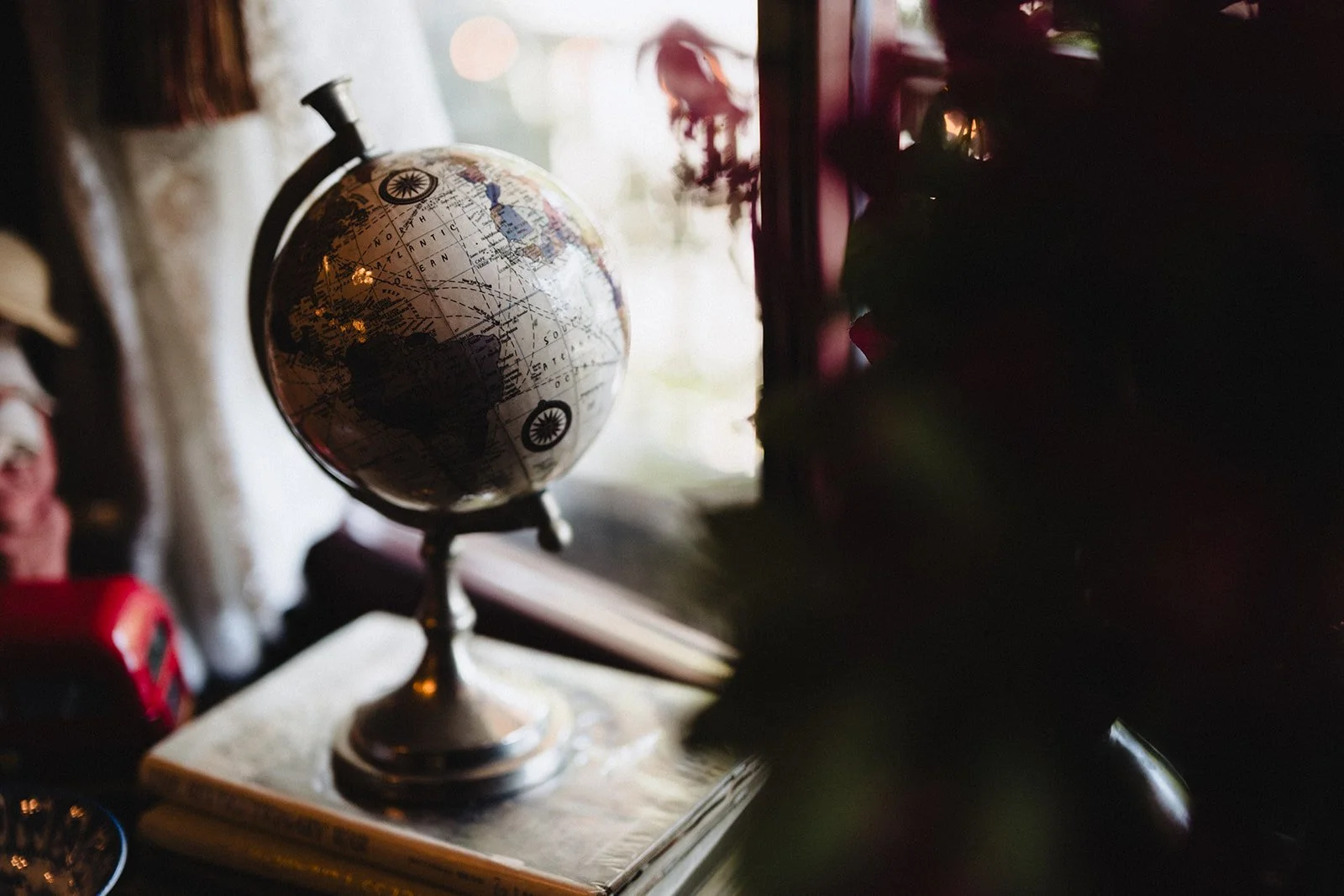 A globe sitting on a table by a window with natural light. The globe is an antique-style world map and is slightly tilted. The background includes objects like a red item and blurred decorations, with a dark shadowed area on the right.