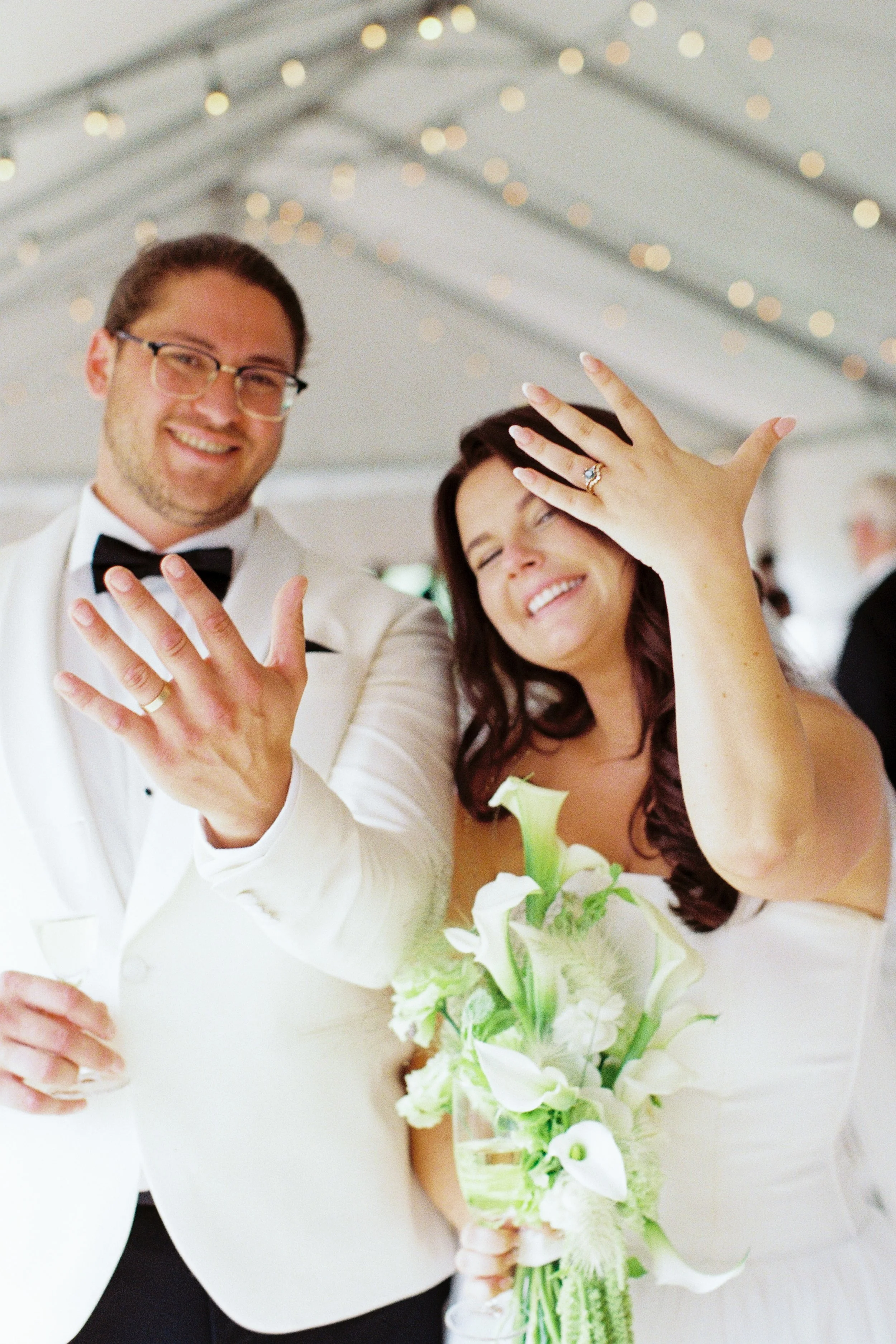 Bride & groom showing off their wedding rings, with the woman holding a bouquet of white flowers.