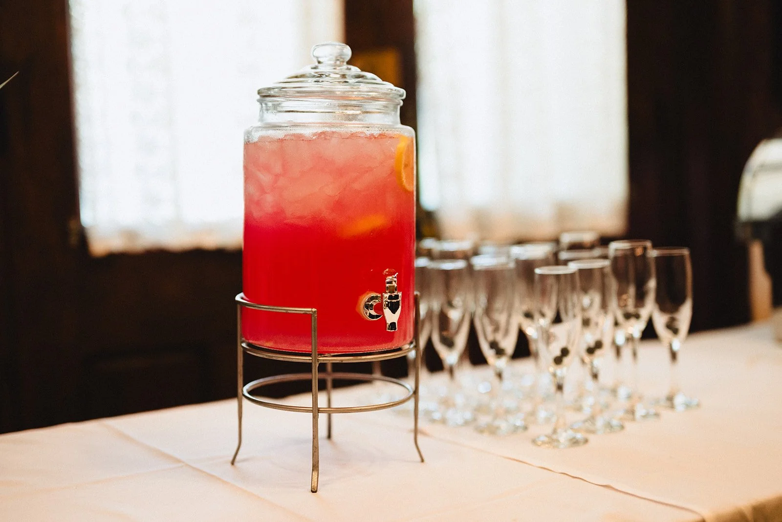 A large glass beverage dispenser with pink-colored drink and ice, with a spout, placed on a metal stand on a table, with empty glasses in the background.
