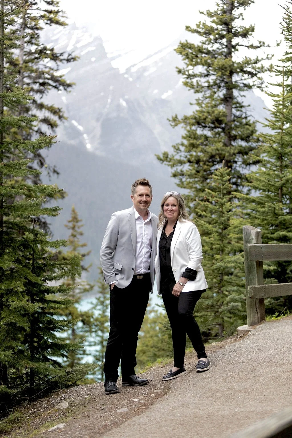 A man and woman smiling outdoors in a mountainous forest area, dressed in business casual attire, with snow-capped peaks in the background.