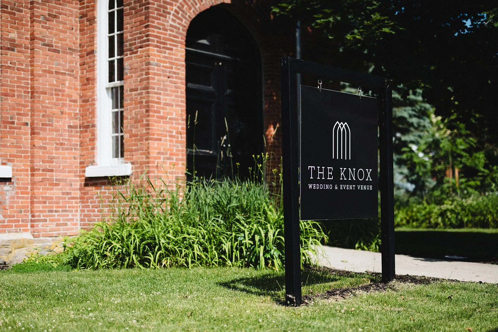 Sign for The Knox wedding and event venue in front of a red brick building with a lawn and lush greenery.