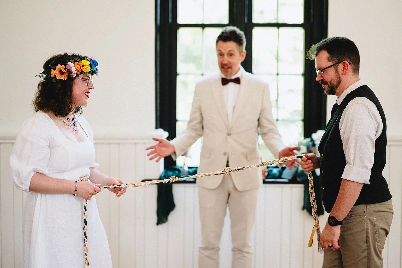 A woman with a flower crown and glasses smiling at a man during a wedding ceremony, holding a rope. The man is looking down at the ropes, dressed in a vest and shirt. An officiant in a cream suit stands in the background, gesturing with his hands in front of a large window.