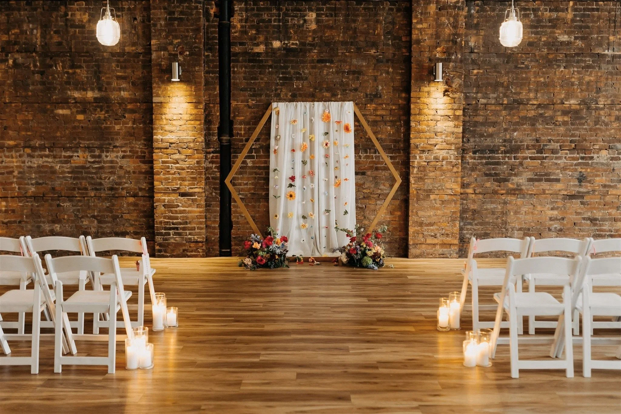 An indoor wedding ceremony setup with white chairs lined up on both sides of a wooden aisle adorned with lit candles. The backdrop features a white curtain decorated with hanging flowers, and the space has exposed brick walls and hanging pendant lights.