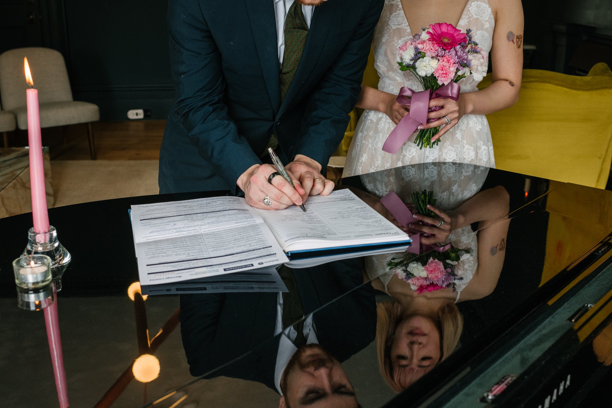 A bride and groom sign the wedding registrar. A pink candle in a glass holder is on the table, and the scene is reflected on a shiny black surface.
