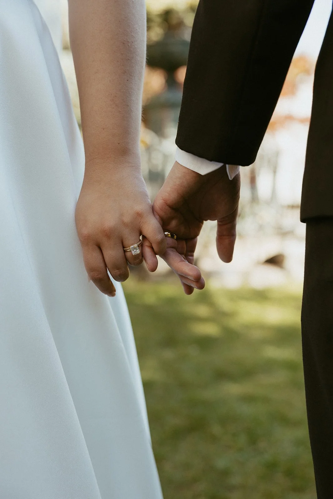 Close-up of a couple holding hands, the woman wearing a white dress and the man in a black suit, with the woman's engagement ring visible.
