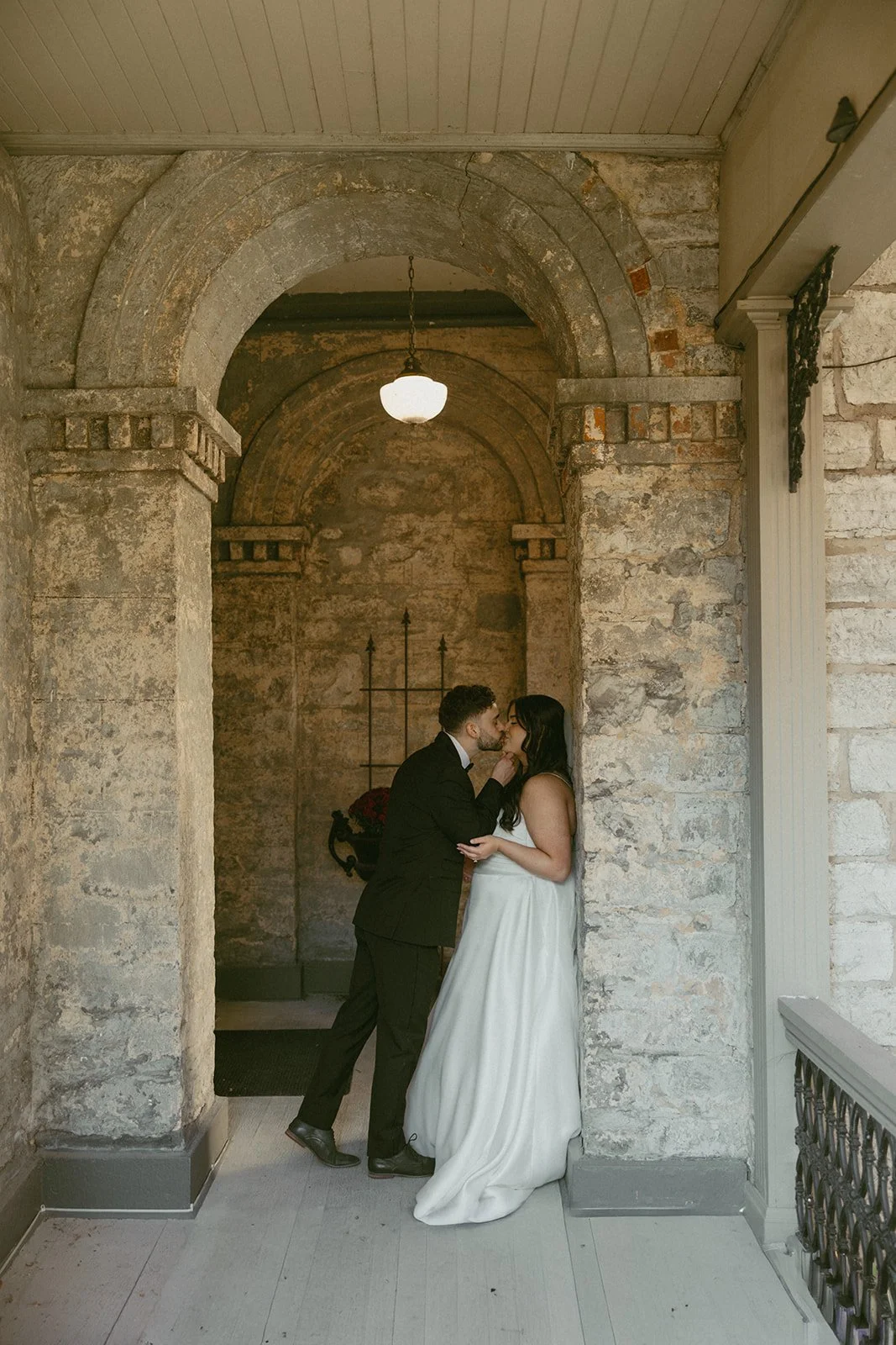 A couple in wedding attire sharing a kiss under an arched stone hallway with rustic brick walls and a hanging lamp.