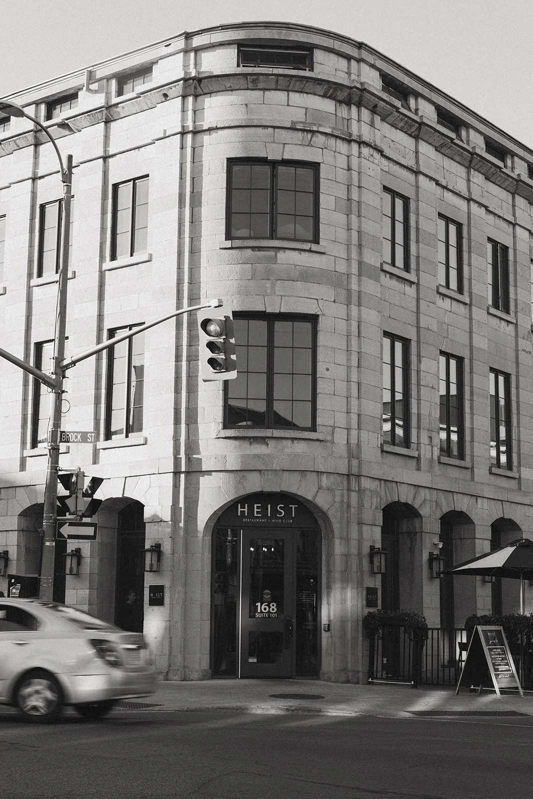 Black and white photo 'HEIST' above the entrance, street sign 'Brock St', traffic light, moving car, and outdoor seating area with an umbrella.