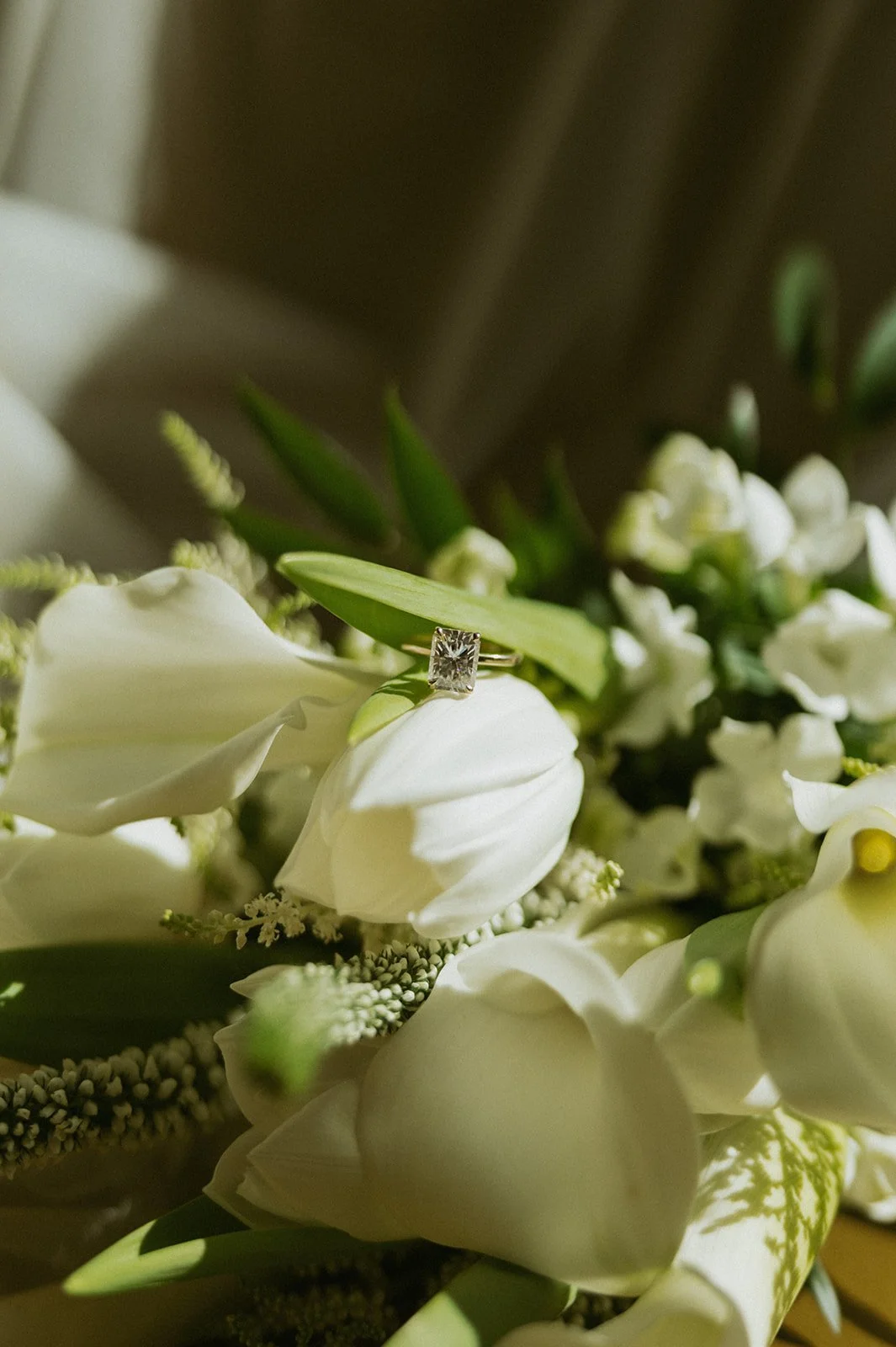 Close-up of a diamond engagement ring resting on white flowers with green leaves.