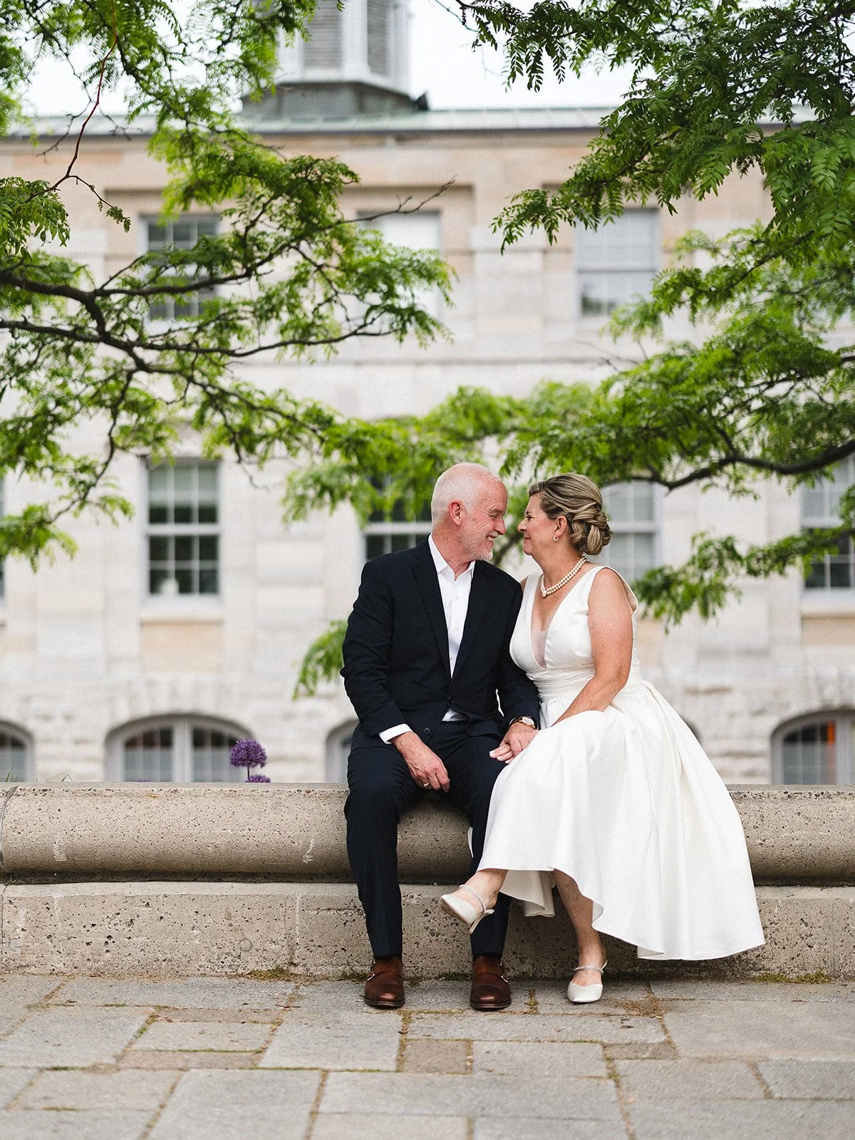 Newlyweds sit closely together on a stone wall. The groom wears a black suit and white shirt, while the bride wears a white dress and pearl necklace. Green trees and a stone building with windows are in the background.