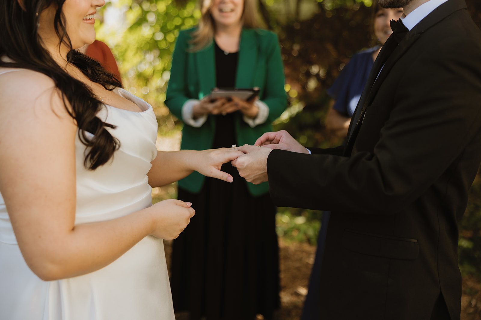 A couple exchanging wedding rings during an outdoor wedding ceremony, with a woman officiant standing behind them, holding a tablet. The bride is wearing a white dress and has dark hair, and the groom is in a black tuxedo.