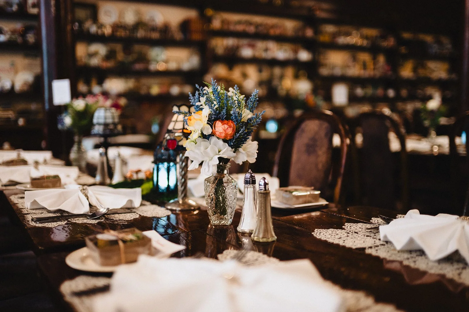 A cozy dining table set with a bouquet of flowers in a glass vase, salt and pepper shakers, and folded napkins with cutlery, in a warmly lit restaurant with wooden chairs and shelves filled with various items in the background.